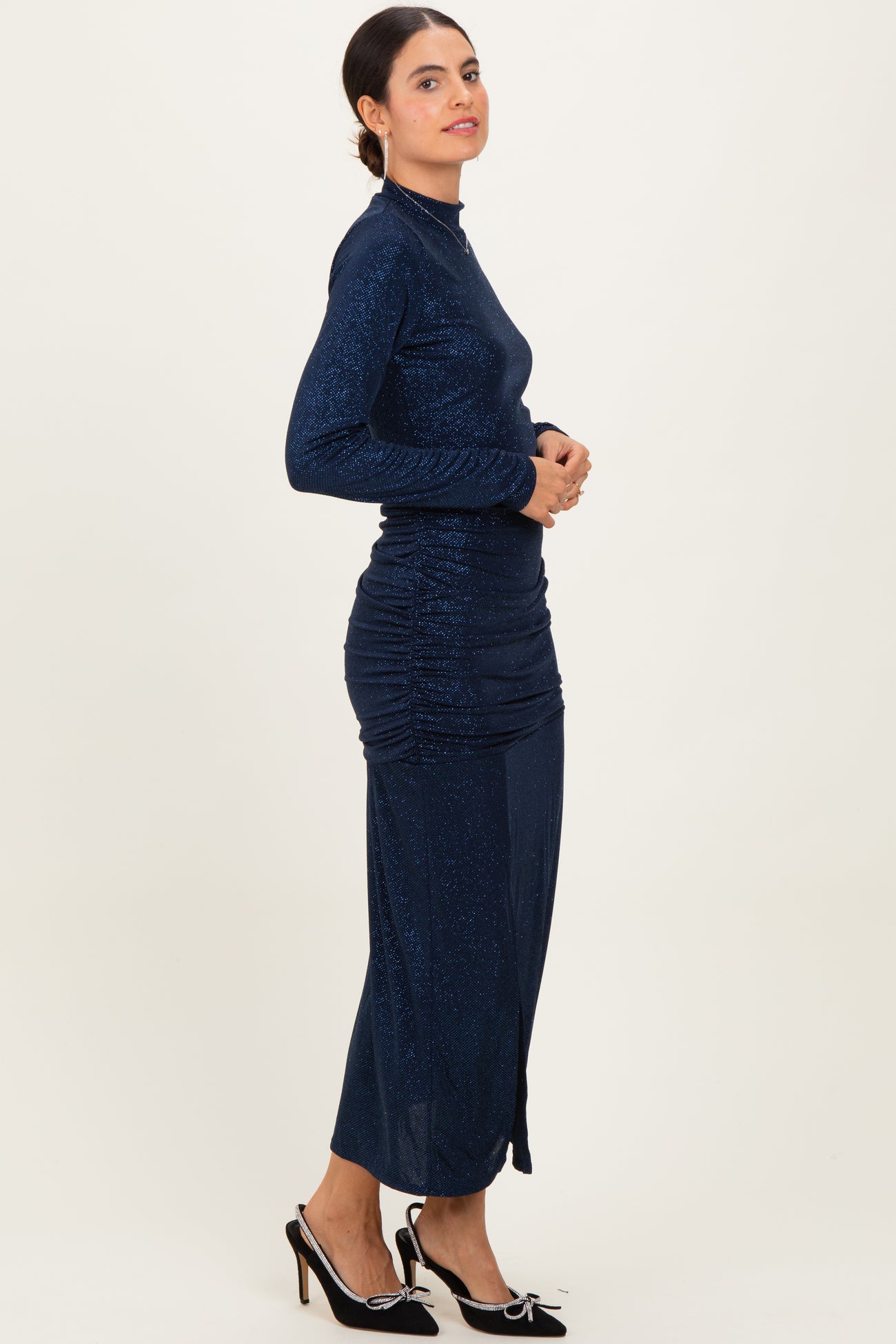 woman wearing a navy blue glitter long sleeve dress with ruching details and a side slit, standing sideways
