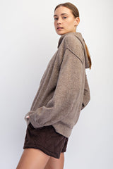 woman wearing a taupe oversized sweater and brown quilted shorts, posing sideways against a plain backdrop