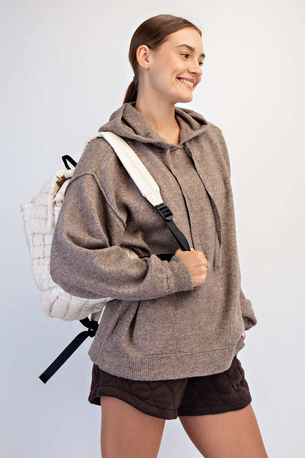 woman wearing a brown oversized hoodie and brown quilted shorts, carrying a cream backpack