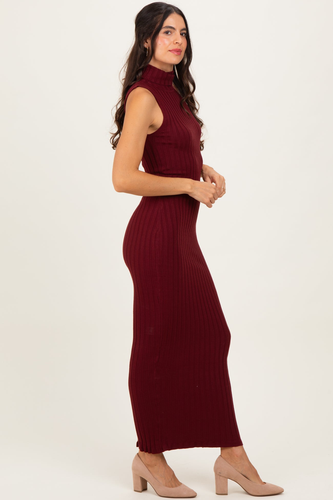 woman wearing a burgundy ribbed knit sleeveless maxi dress and beige heels, standing sideways against a neutral background