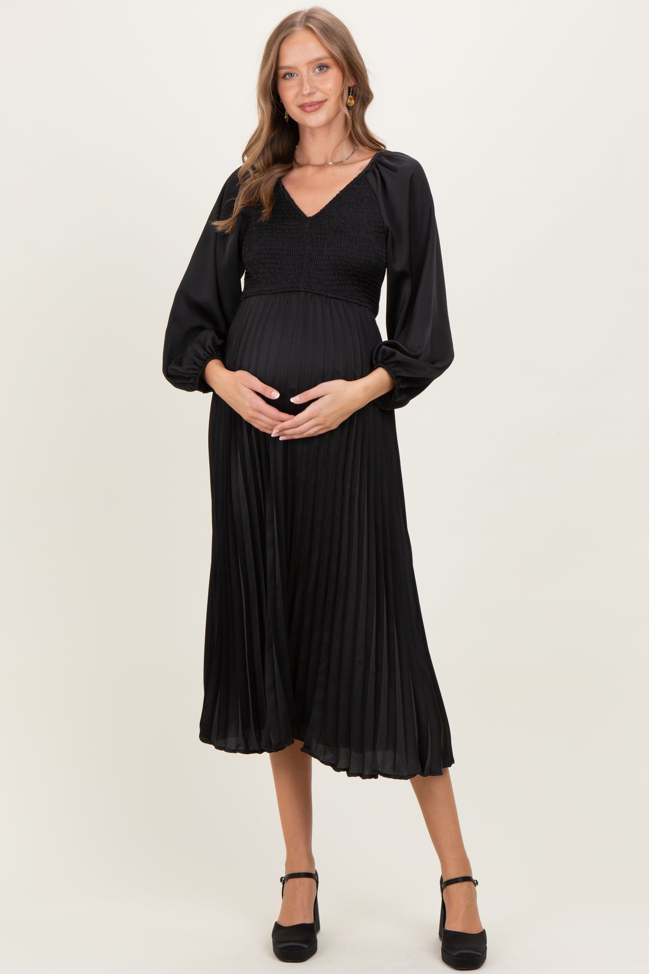 woman wearing a black pleated maternity dress with a smocked bodice and balloon sleeves, holding her baby bump