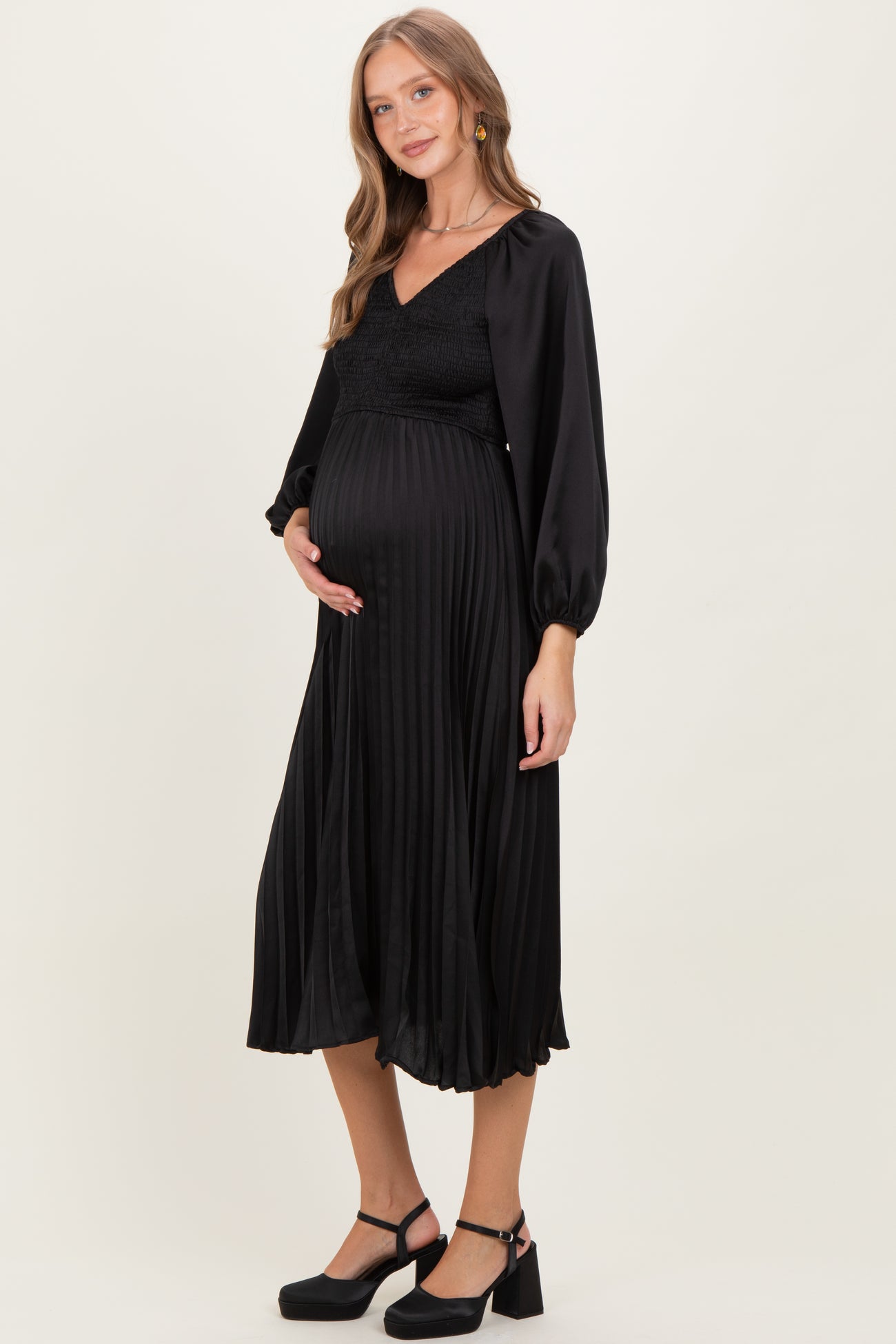 woman wearing a black pleated maternity dress with long sleeves and a fitted bodice, holding her baby bump