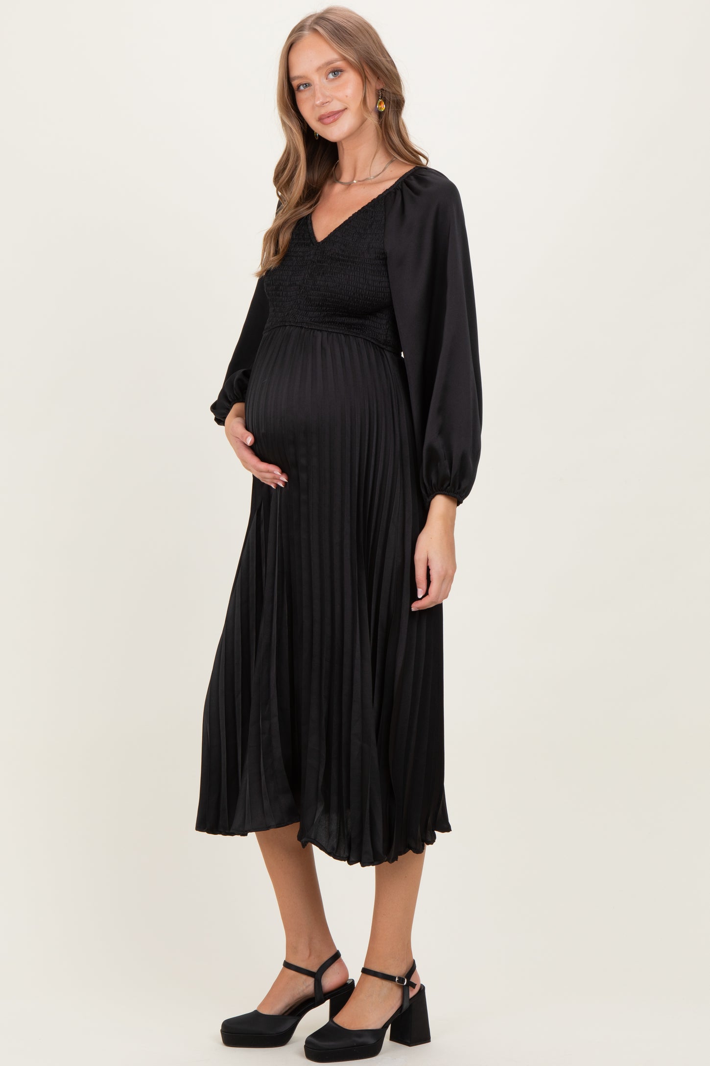 woman wearing a black pleated maternity dress with long sleeves and a fitted bodice, holding her baby bump