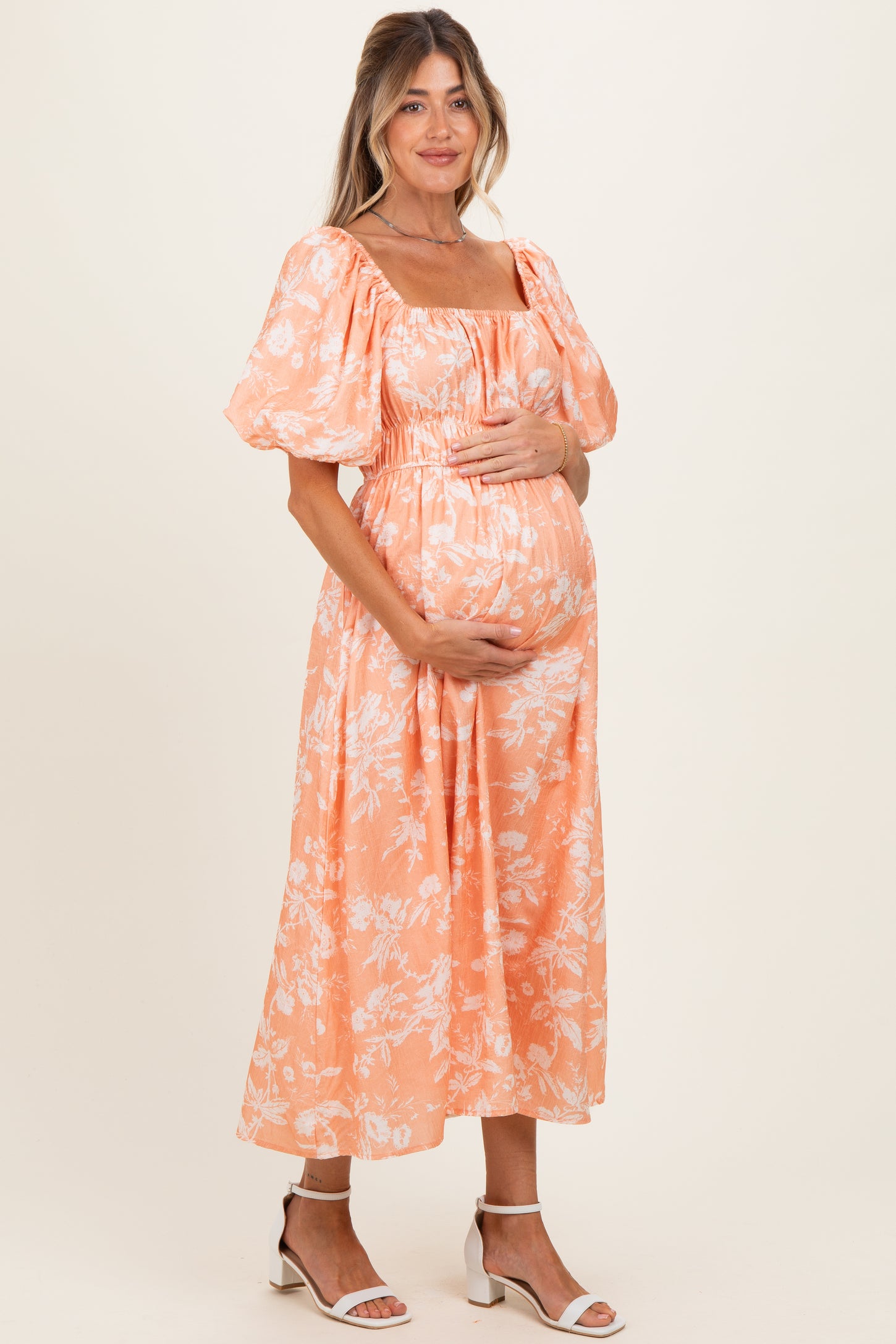 woman wearing an apricot floral maternity maxi dress, holding her baby bump with a smile
