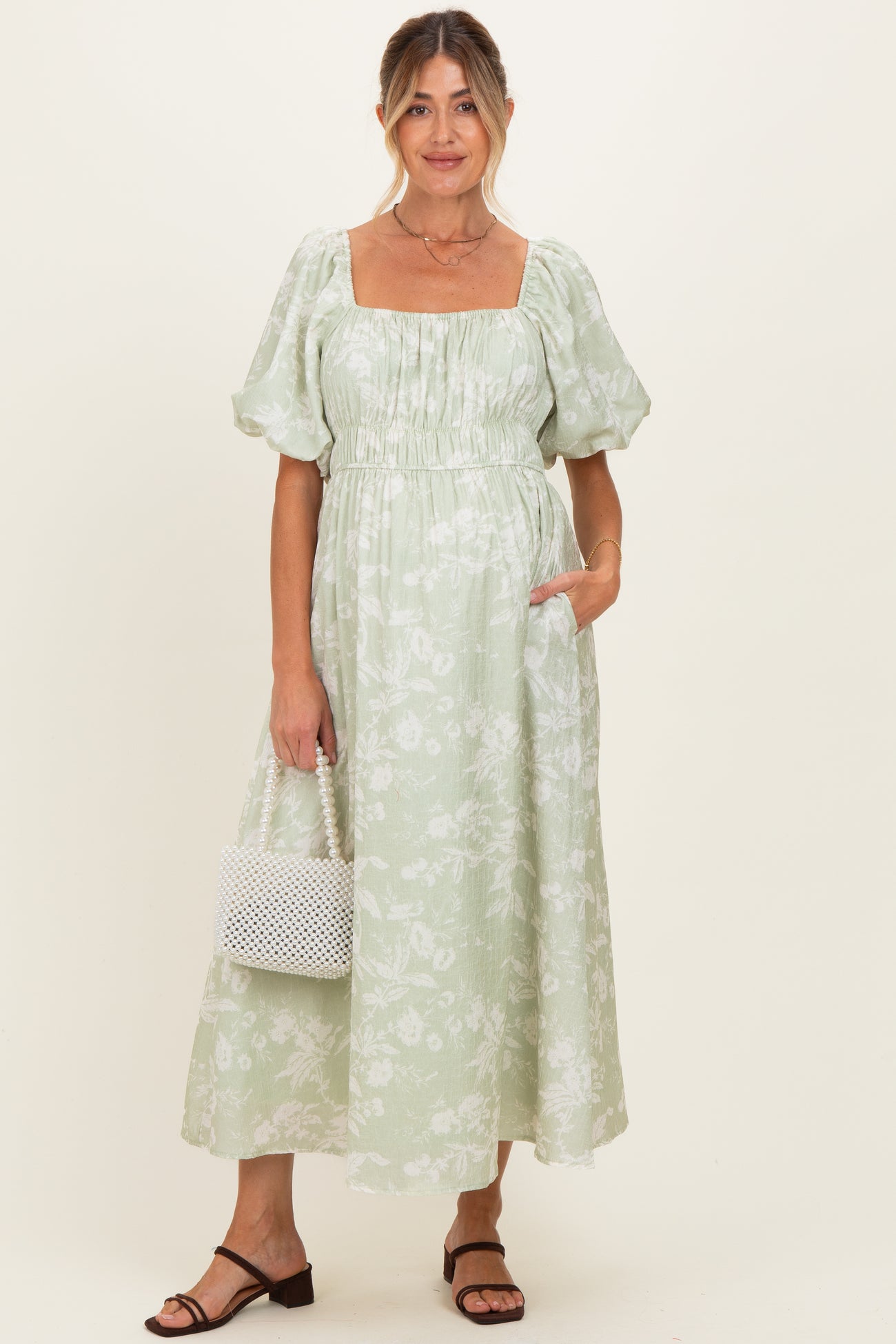 woman wearing a light green floral maternity dress with puffed sleeves and pockets, holding a white bag