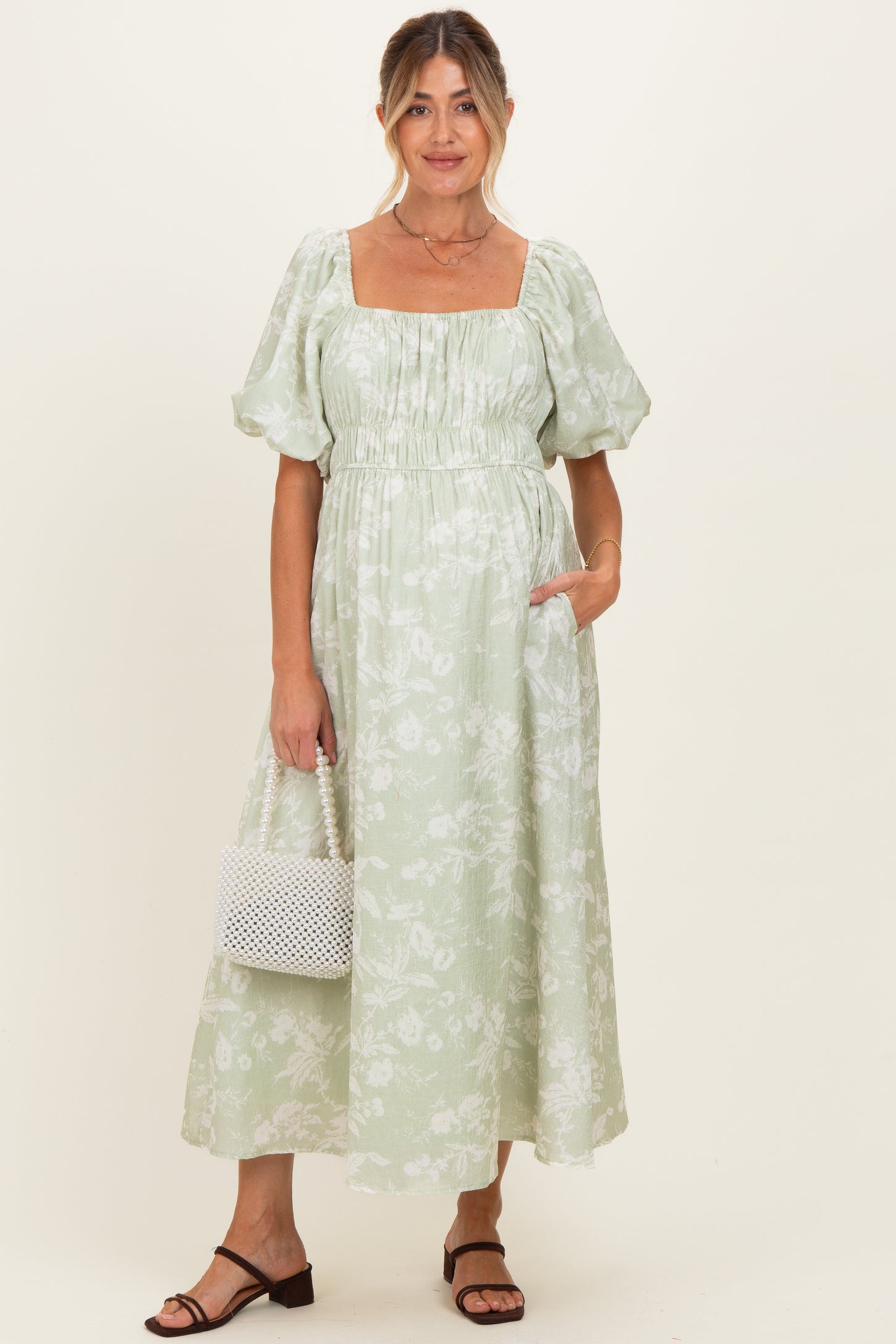 woman wearing a light green floral maternity dress with puffed sleeves and pockets, holding a white bag