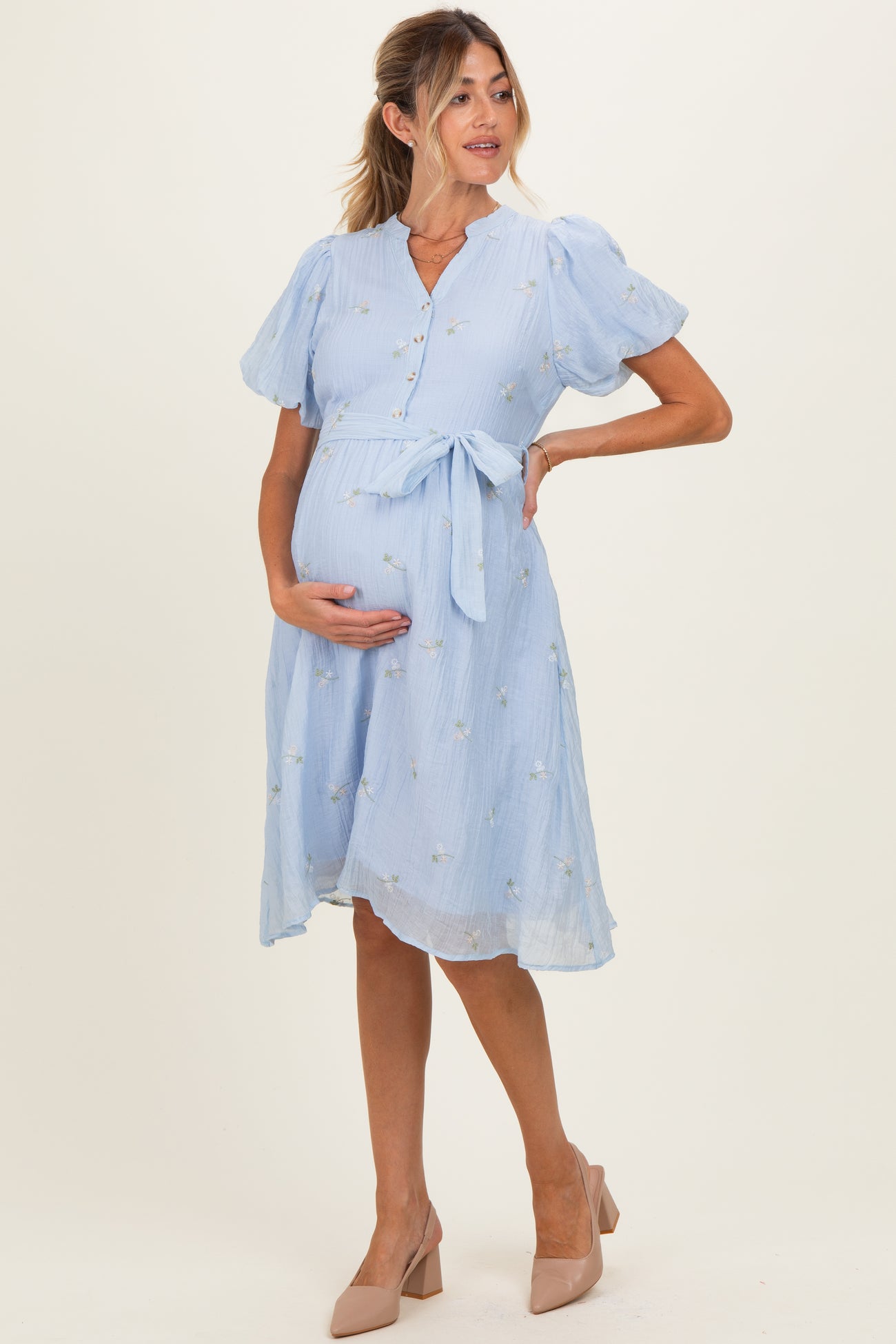 woman wearing a light blue maternity dress with puff sleeves and floral details, holding her baby bump