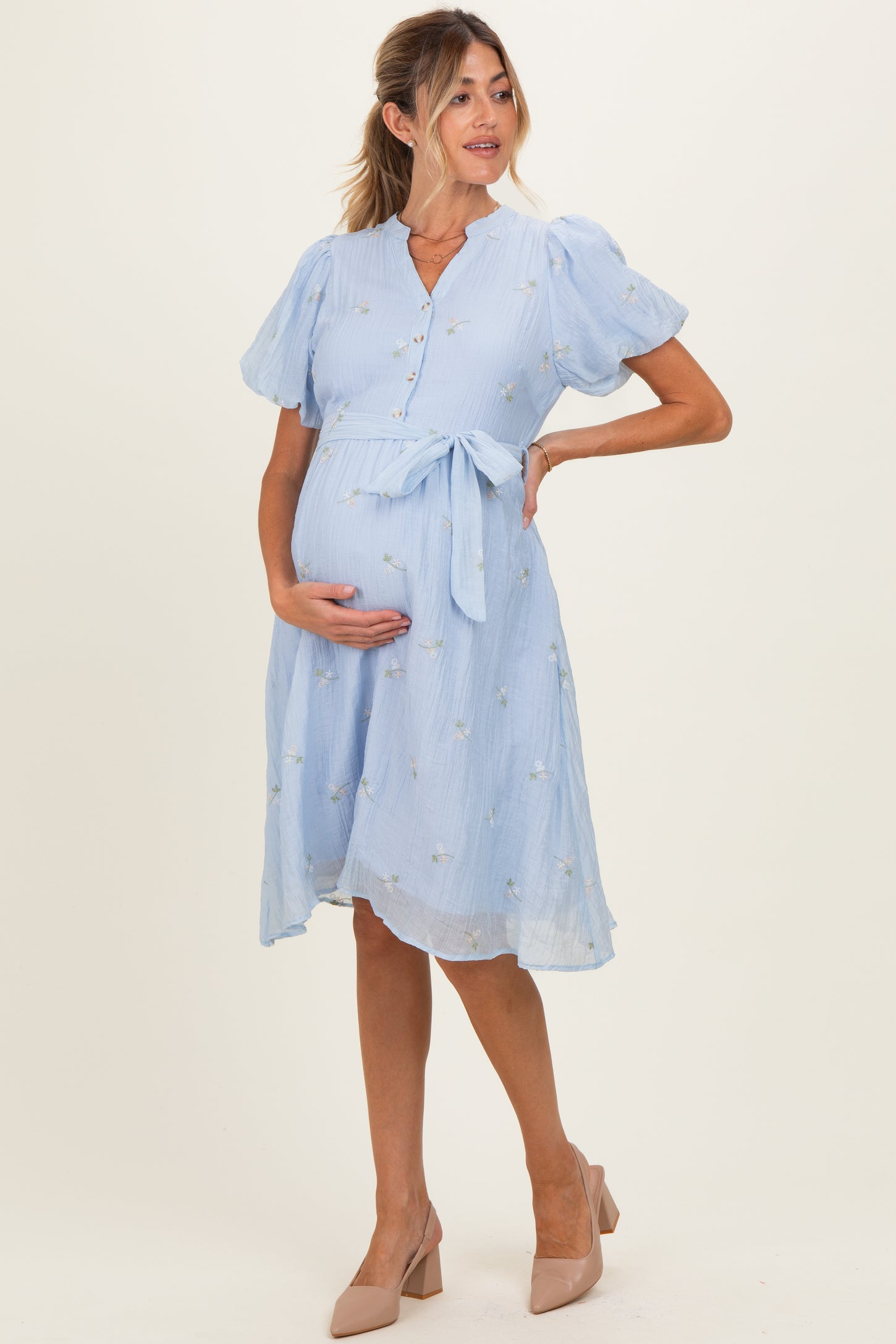woman wearing a light blue maternity dress with puff sleeves and floral details, holding her baby bump