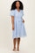 woman wearing a light blue puff sleeve dress with floral embroidery and a tied waist, standing with her hands in pockets