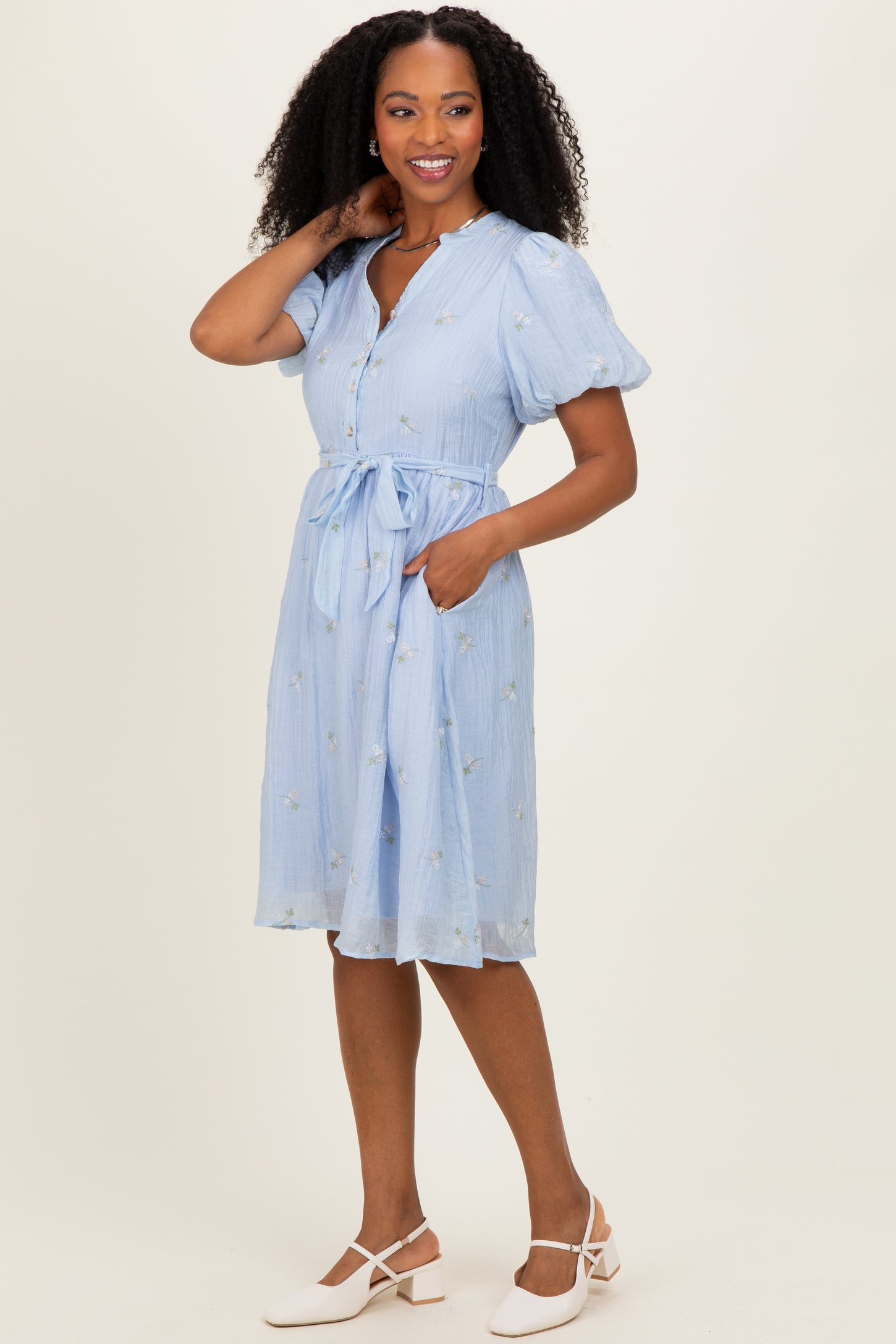 woman wearing a light blue short sleeve dress with a tie waist and floral embroidery, standing with one hand in the pocket