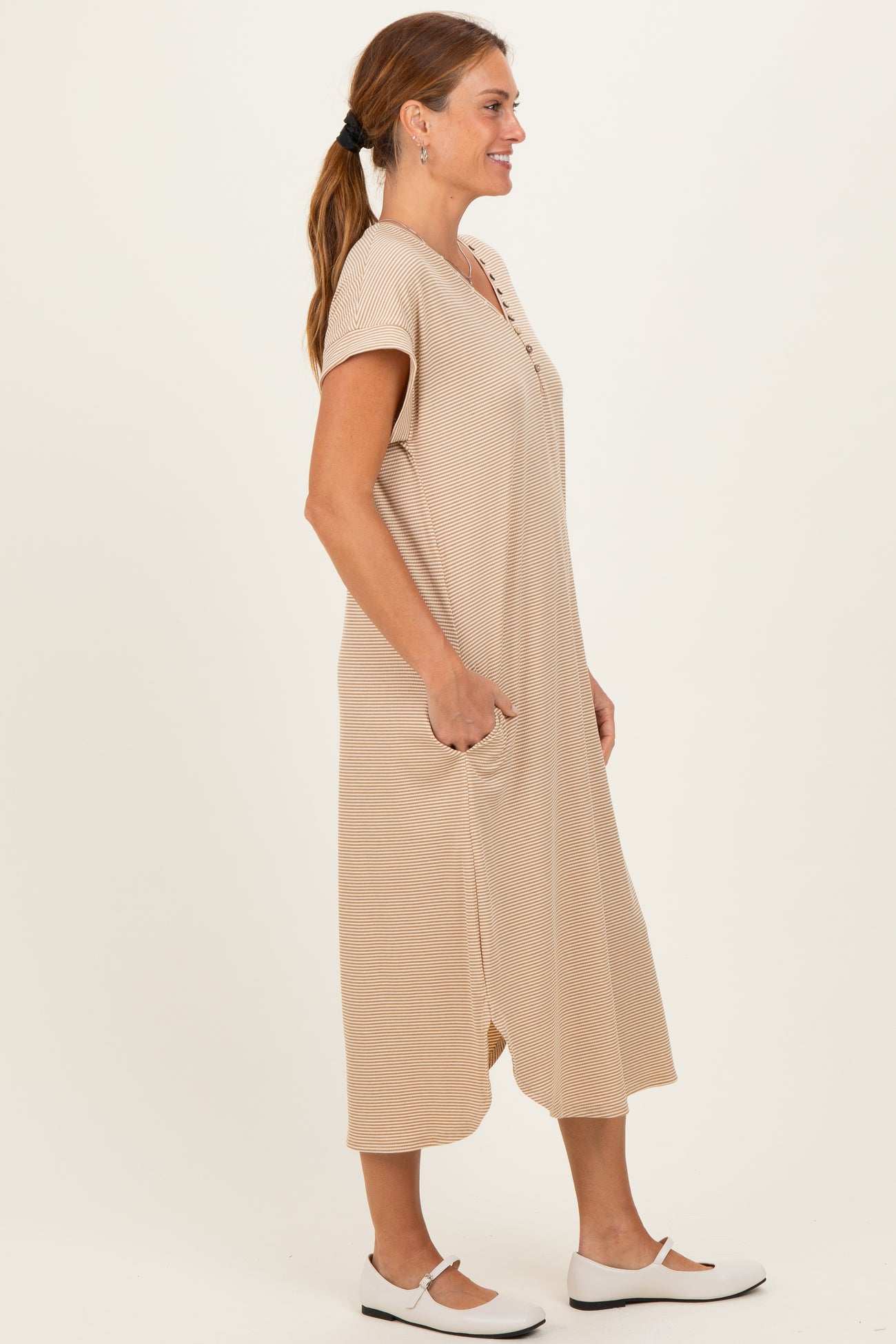 woman wearing a beige short sleeve midi dress with buttons and pockets, standing sideways against a neutral background