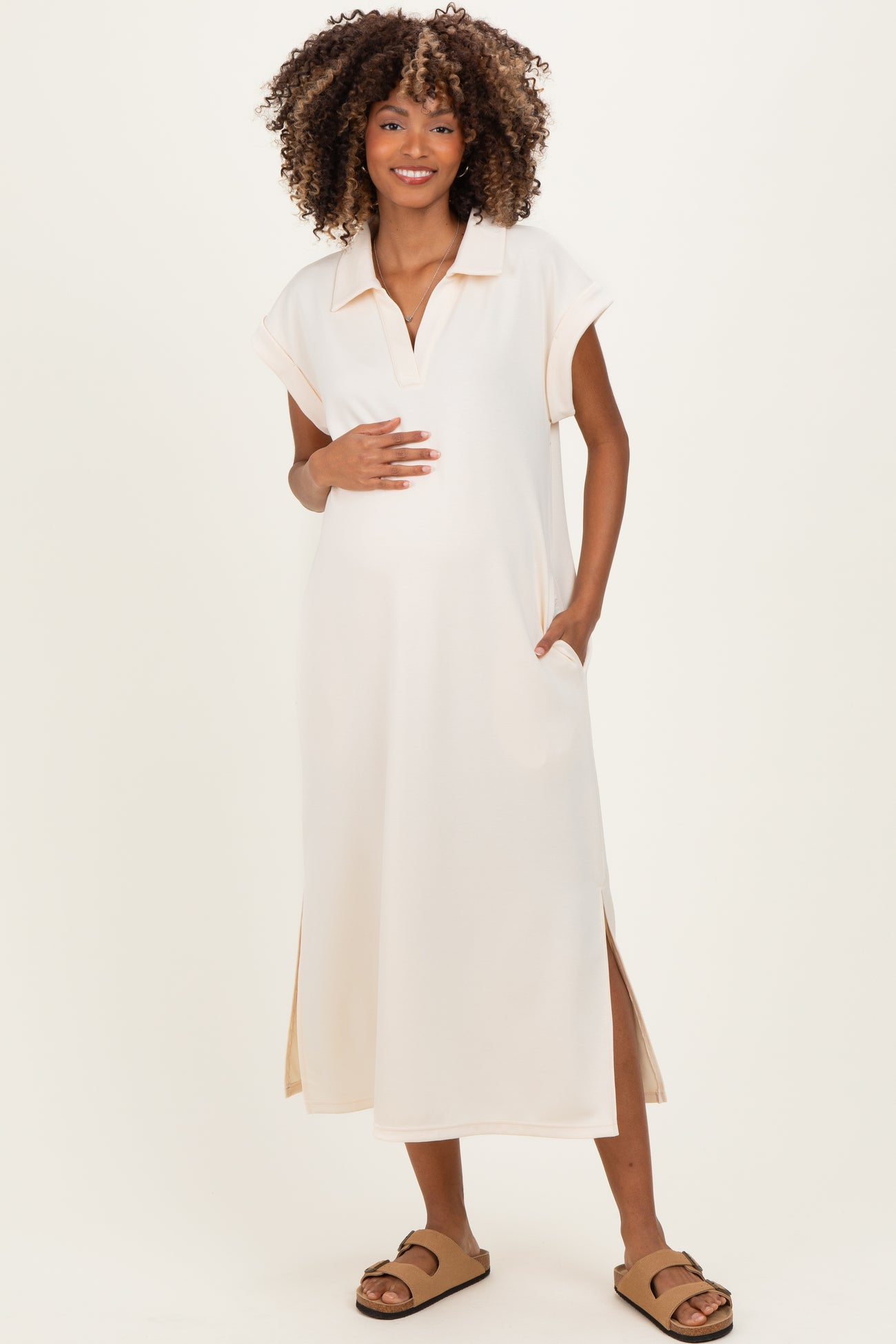 woman wearing a cream short sleeve maternity dress with a collar and side slits, holding her baby bump