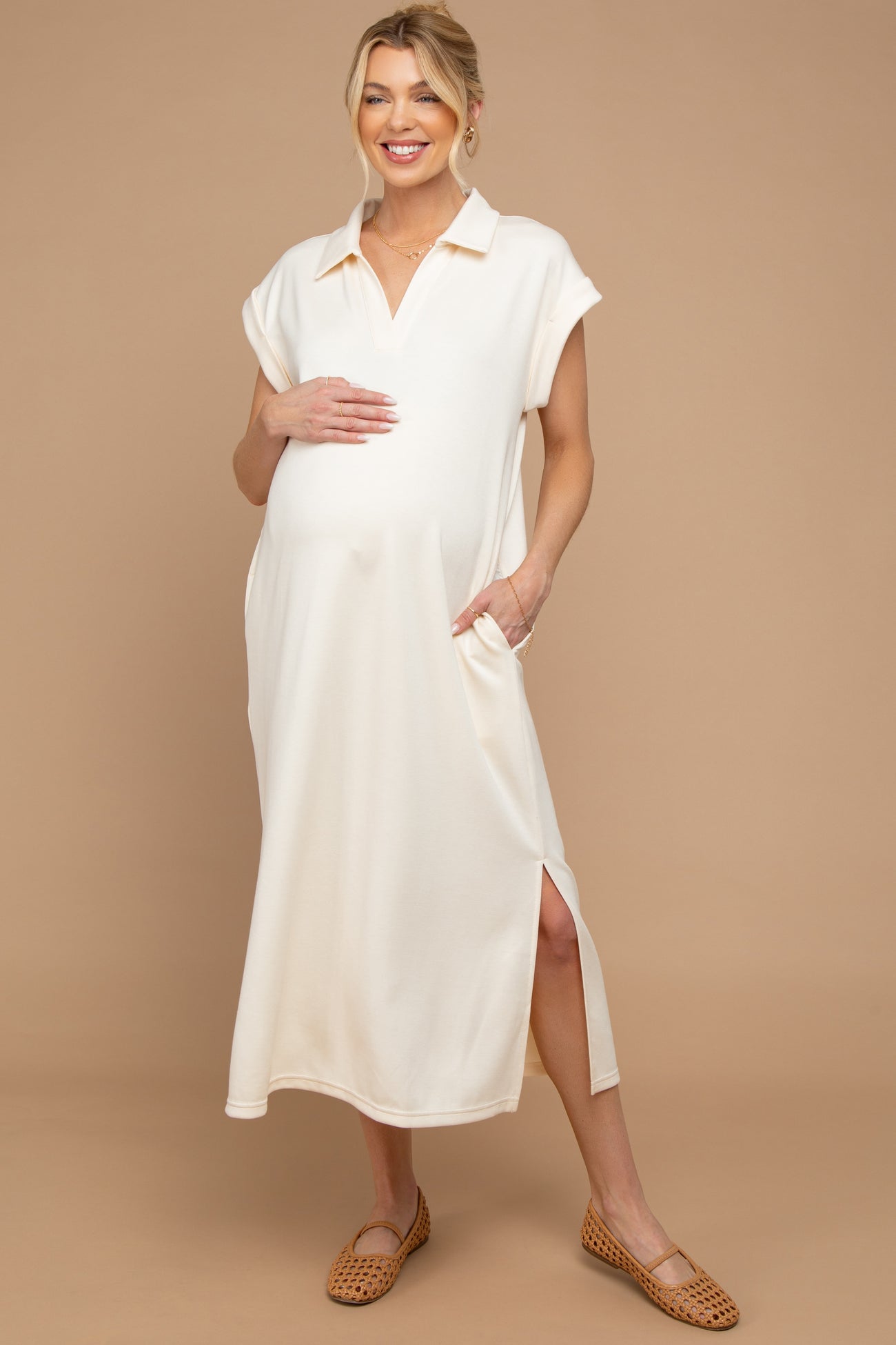 woman wearing a cream maternity dress with short sleeves, pockets, and side slits, cradling her baby bump