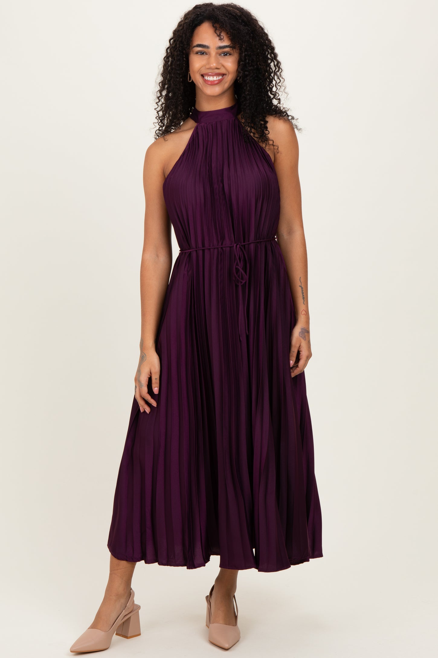 woman wearing a sleeveless deep purple pleated maxi dress with a halter neck and tie waist detail