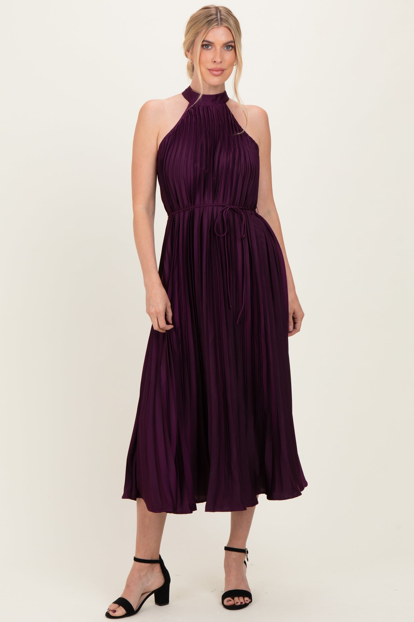 woman wearing a sleeveless plum pleated midi dress with a halter neckline and a tied waist detail