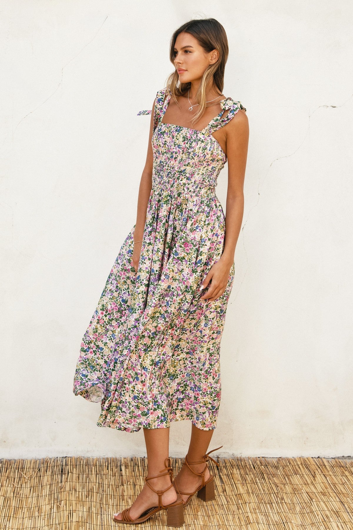 woman wearing a floral midi dress with shoulder ties and a smocked bodice, standing against a textured wall