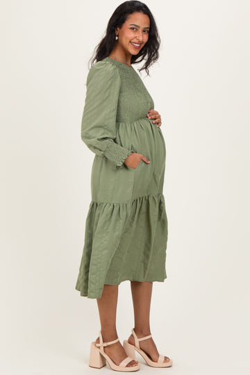 woman wearing a long sleeve sage green maternity dress with a smocked bodice and tiered skirt, showing her baby bump