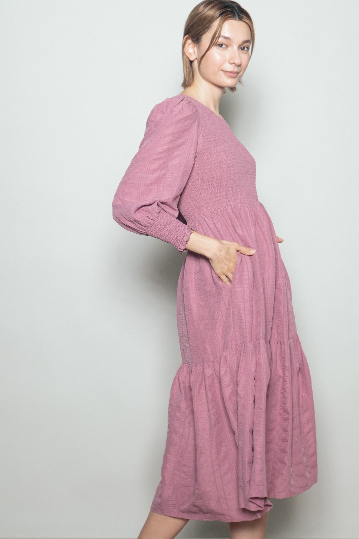 woman wearing a mauve long sleeve midi dress with a smocked bodice and tiered skirt, featuring side pockets