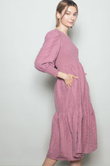 woman wearing a mauve long sleeve midi dress with a smocked bodice and tiered skirt, featuring side pockets