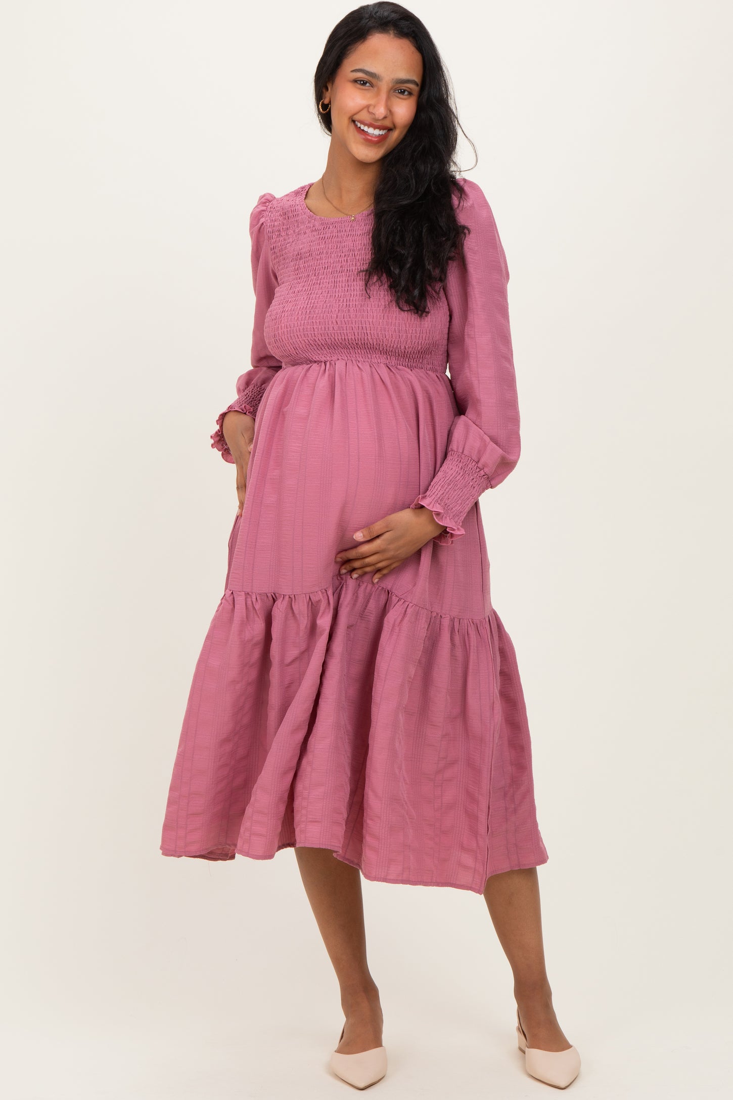 woman wearing a pink maternity dress with long sleeves, smocked bodice, and tiered skirt holding her baby bump