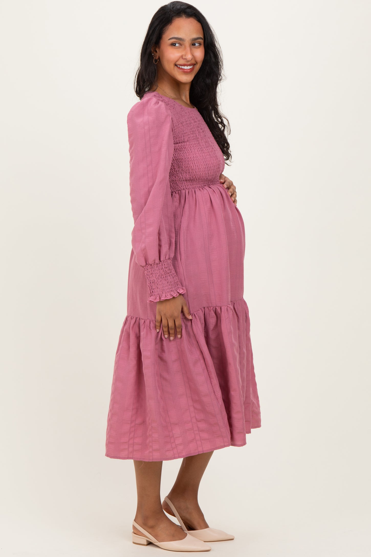 woman wearing a pink long sleeve maternity dress with smocked bodice and tiered skirt, showcasing her baby bump