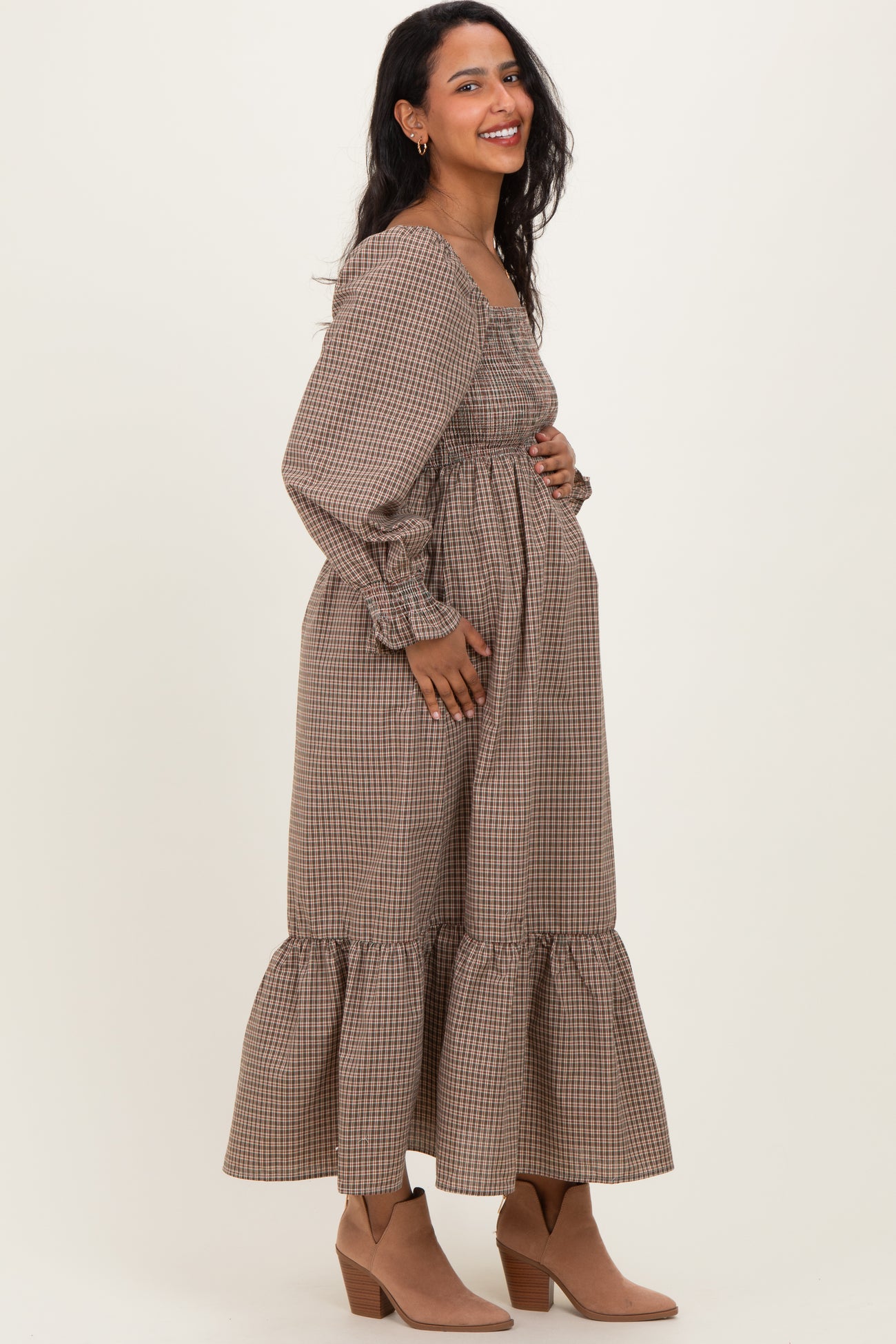 woman wearing a brown gingham long sleeve maternity dress with a tiered skirt and holding her baby bump