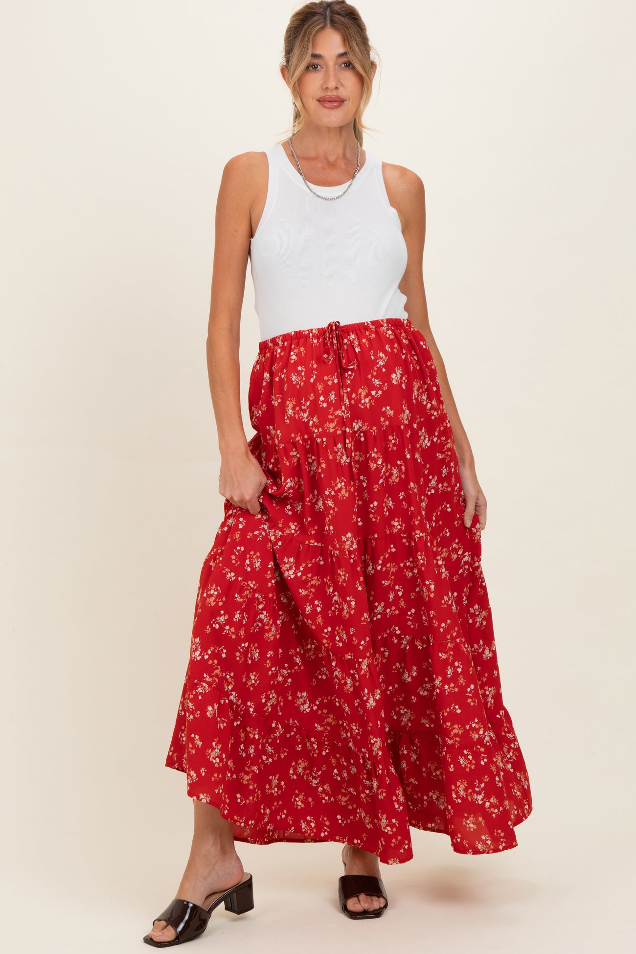 woman wearing a white tank top and a long red floral maternity skirt, gently holding her baby bump