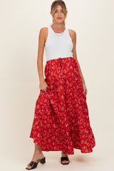 woman wearing a white tank top and a long red floral maternity skirt, gently holding her baby bump