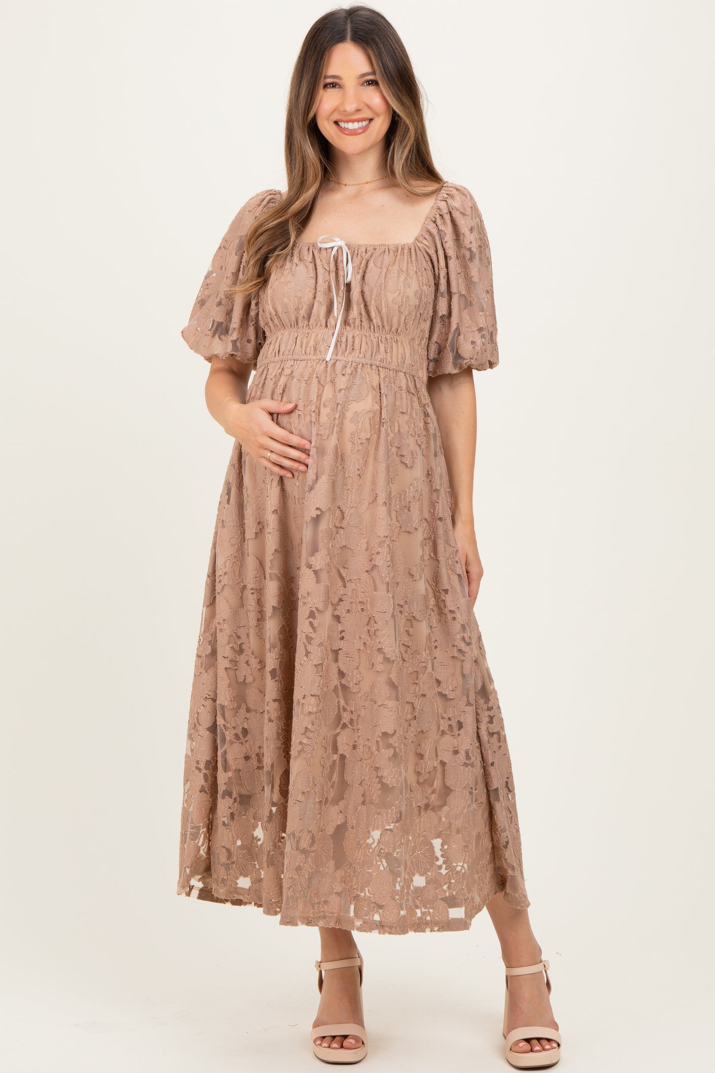 woman wearing a blush lace maternity dress with puff sleeves and a tie detail, holding her baby bump