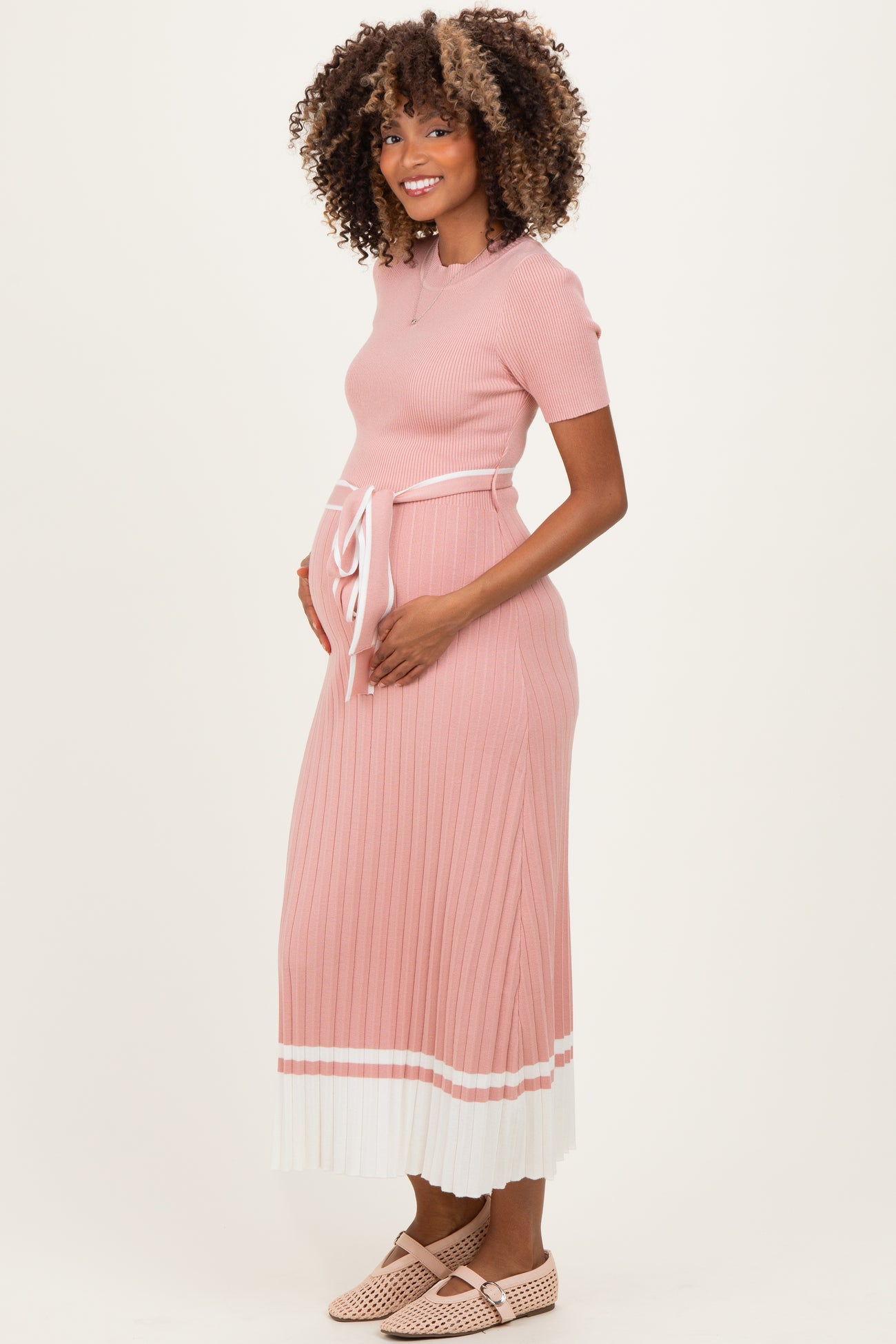 woman wearing a pink ribbed maternity dress with short sleeves, a tie waist, and a pleated skirt, holding her baby bump
