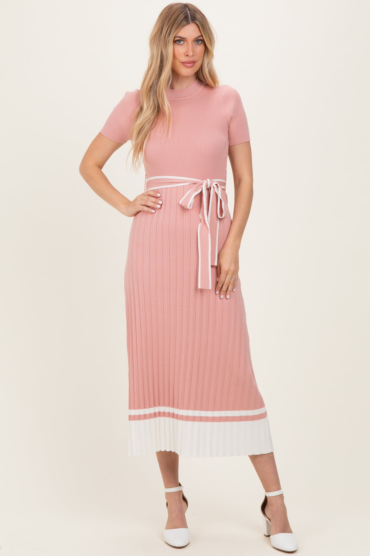 woman wearing a pink ribbed midi dress with a white tie waist and contrasting hem details, standing against a neutral background