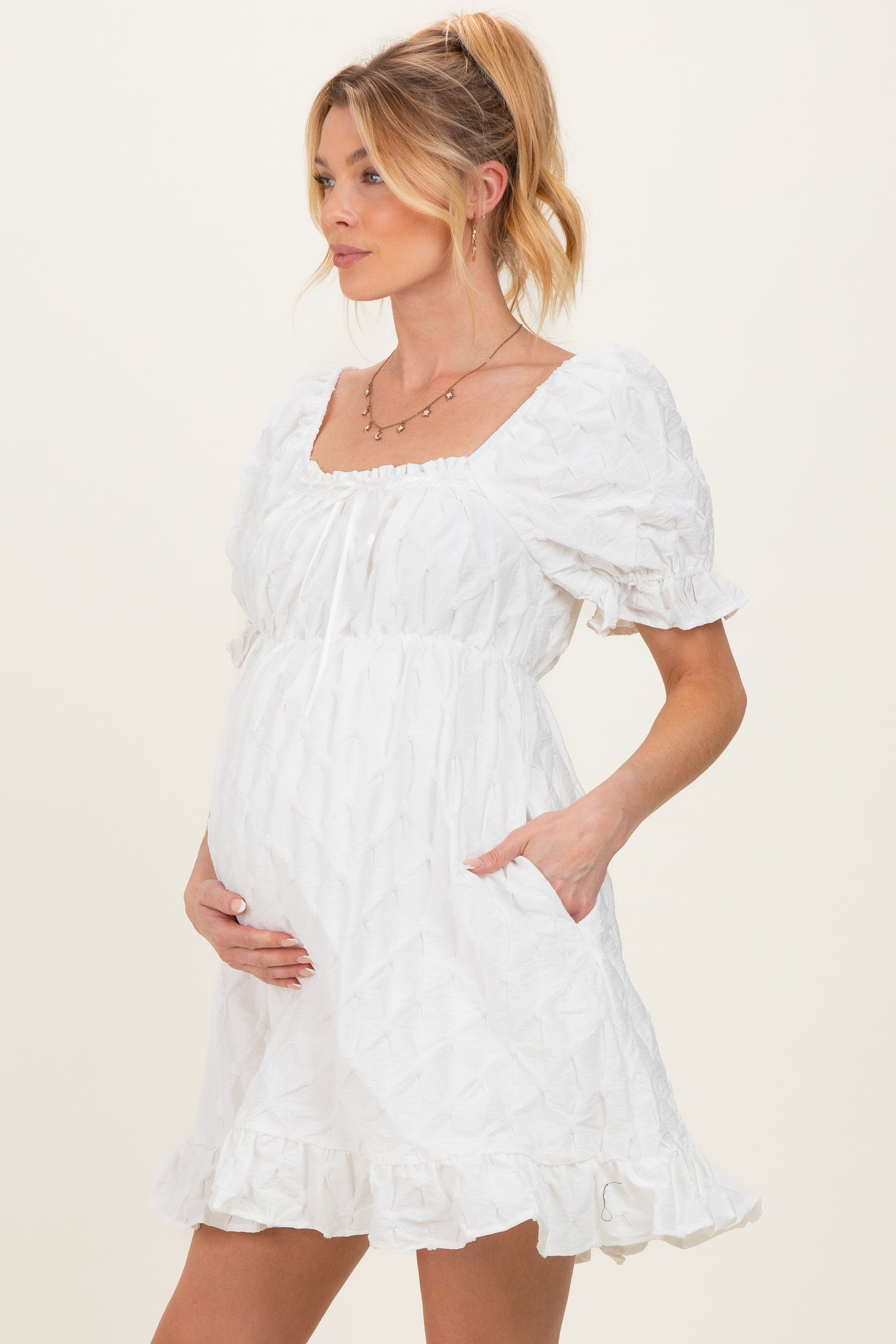woman wearing a white short puff sleeve maternity dress with a square neckline and pockets, holding her baby bump