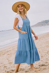 woman wearing a light blue tiered maxi dress with pockets and a white tee, standing on the beach with a straw hat