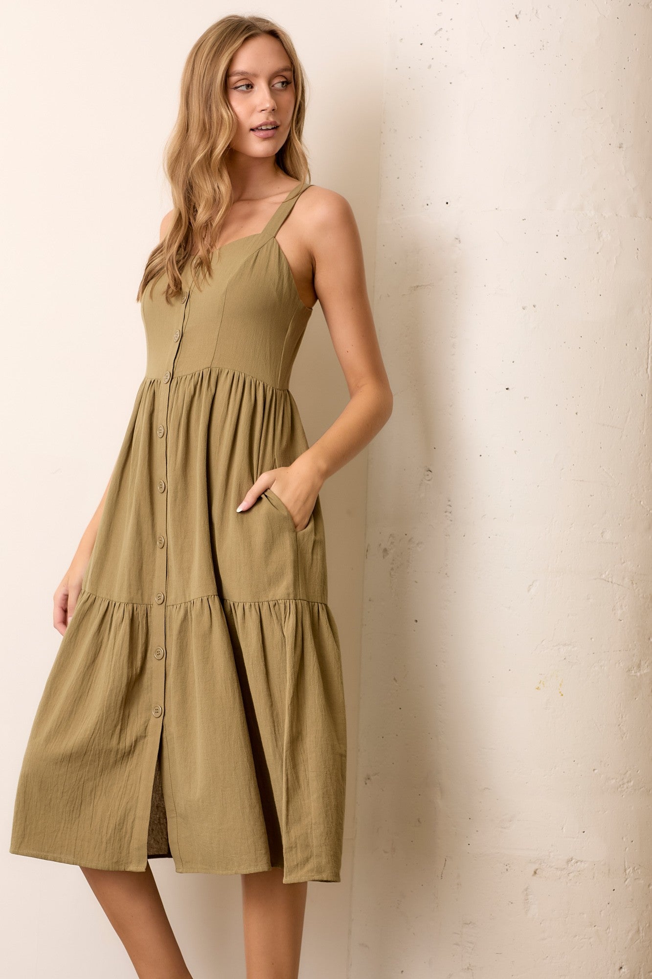 woman wearing a khaki green midi dress with button details and pockets, featuring spaghetti straps and a tiered skirt.