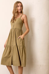 woman wearing a khaki green midi dress with button details and pockets, featuring spaghetti straps and a tiered skirt.