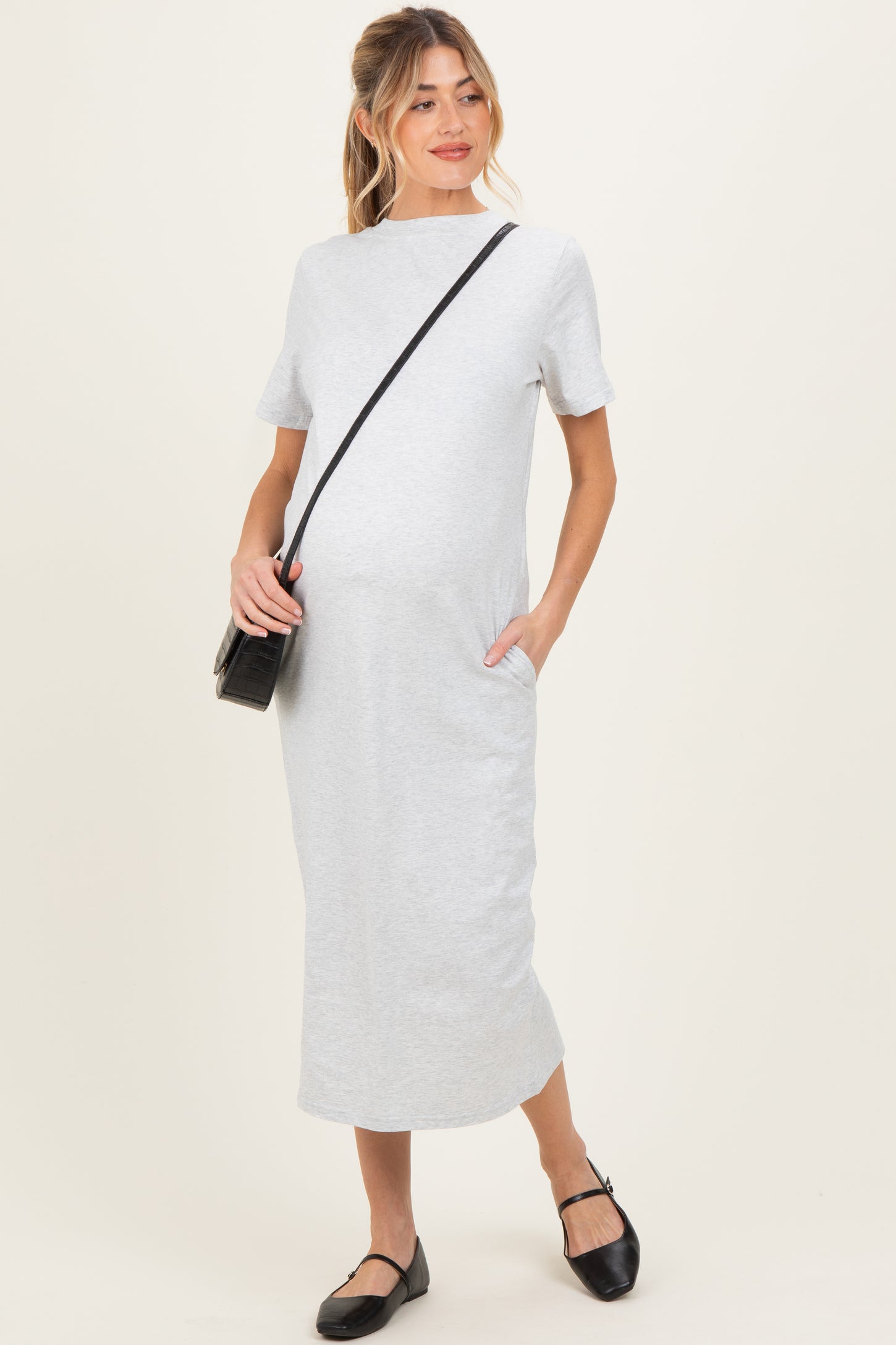 woman wearing a gray maternity dress with short sleeves and pockets, holding a black crossbody bag and showing a baby bump