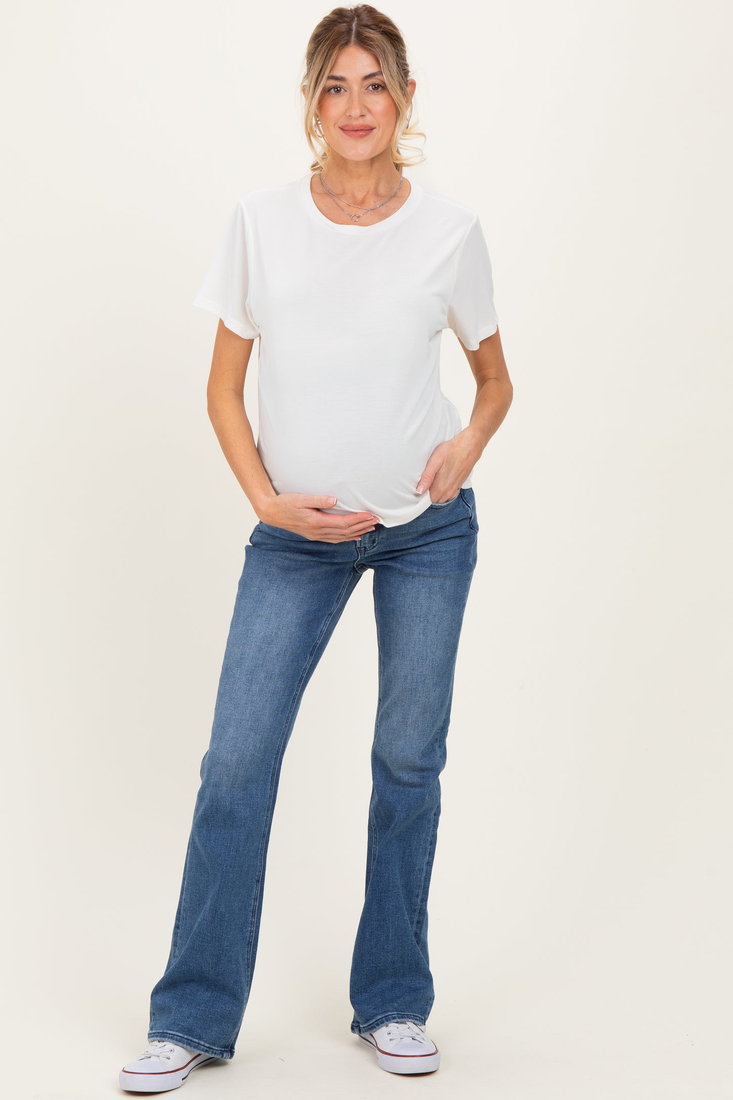 woman wearing a white short sleeve maternity top and blue flared jeans, holding her baby bump