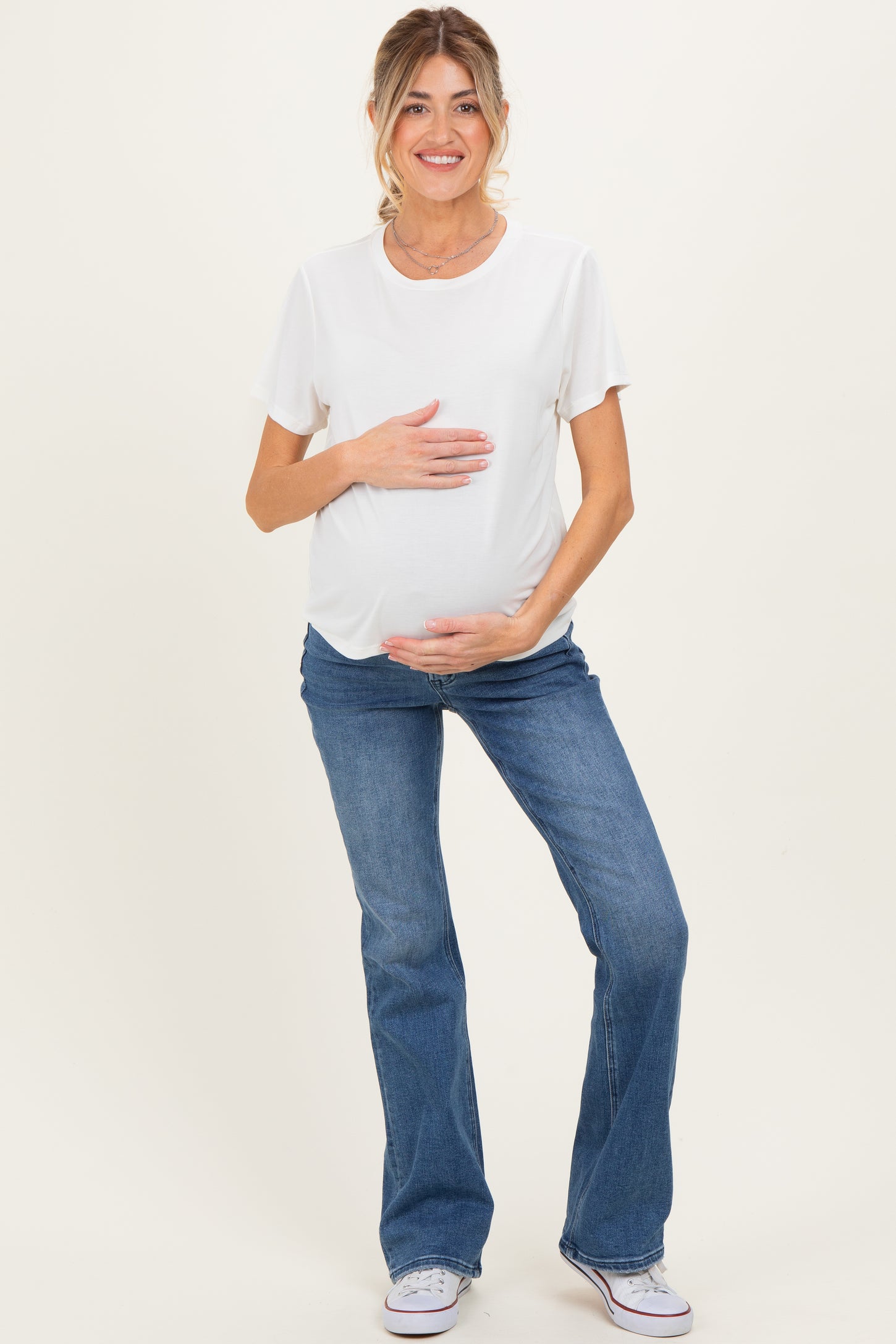 woman wearing a white short sleeve maternity top and blue flared maternity jeans, holding her baby bump