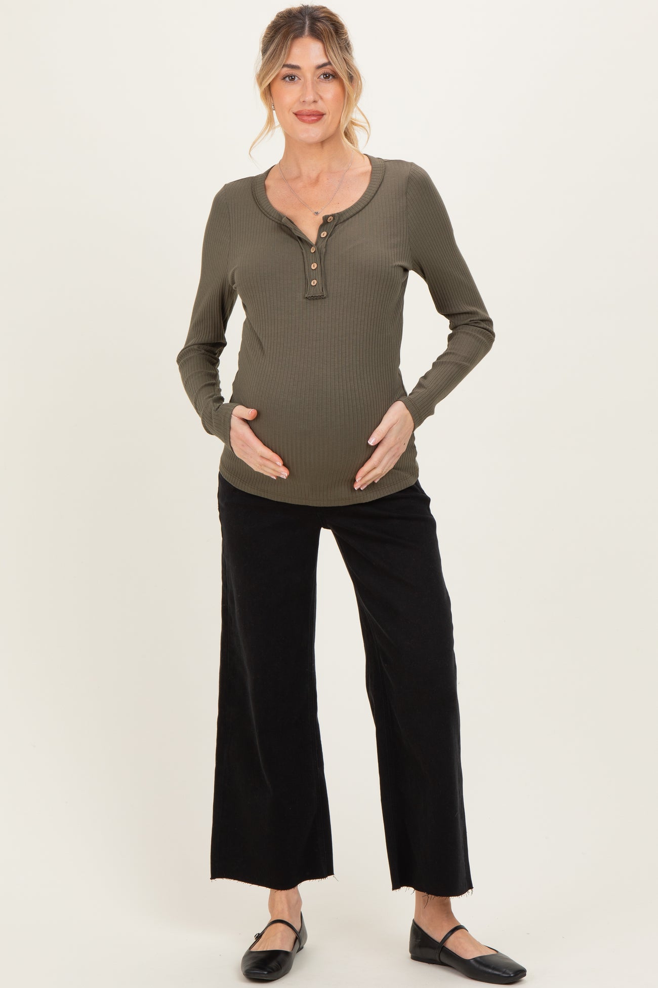 woman wearing a dark green long sleeve maternity top with buttons and black wide-leg pants, gently cradling her baby bump