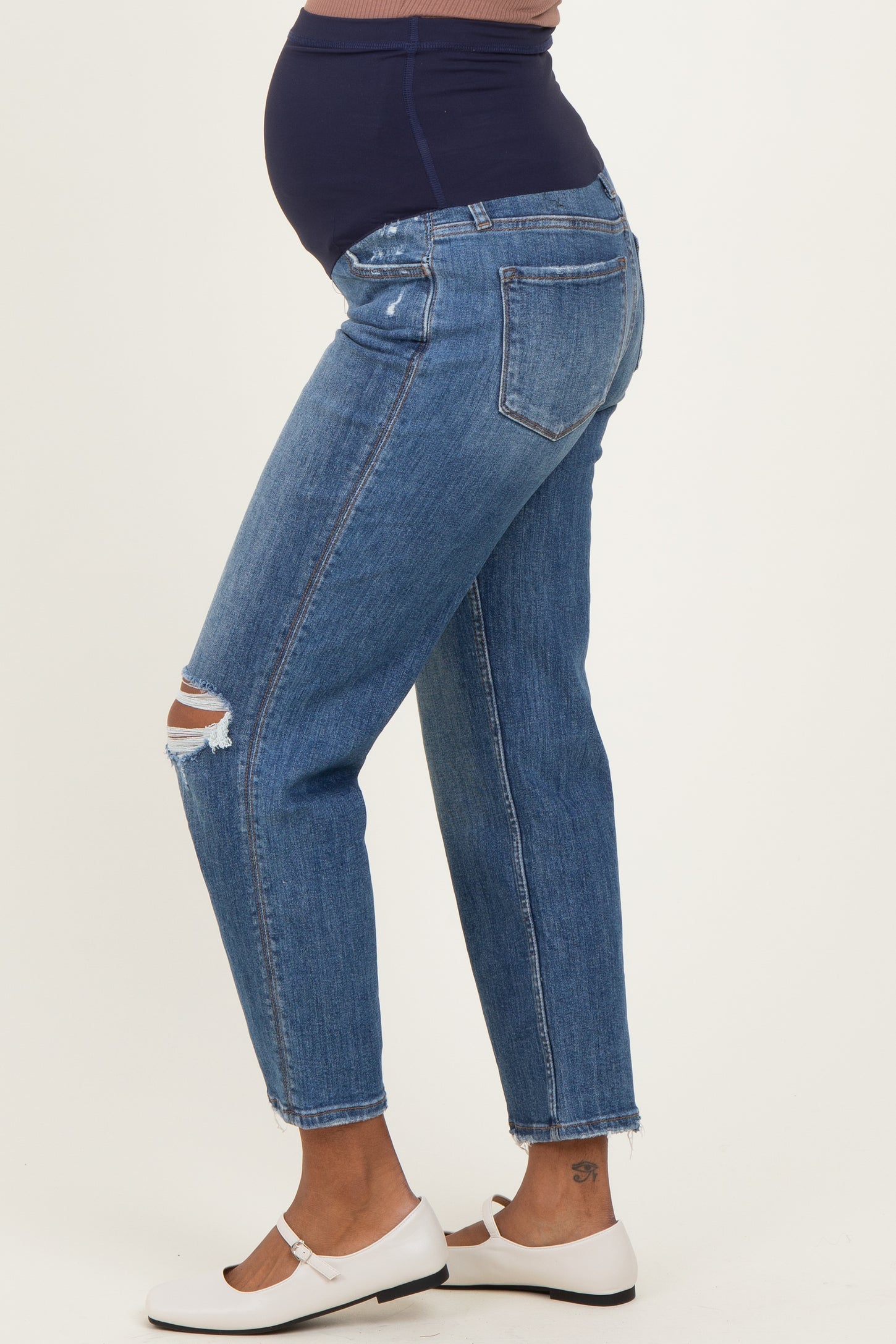 woman wearing blue maternity jeans with a visible baby bump and distressed details, styled with white flats