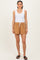 woman wearing a white tank top and brown drawstring shorts, standing against a neutral background