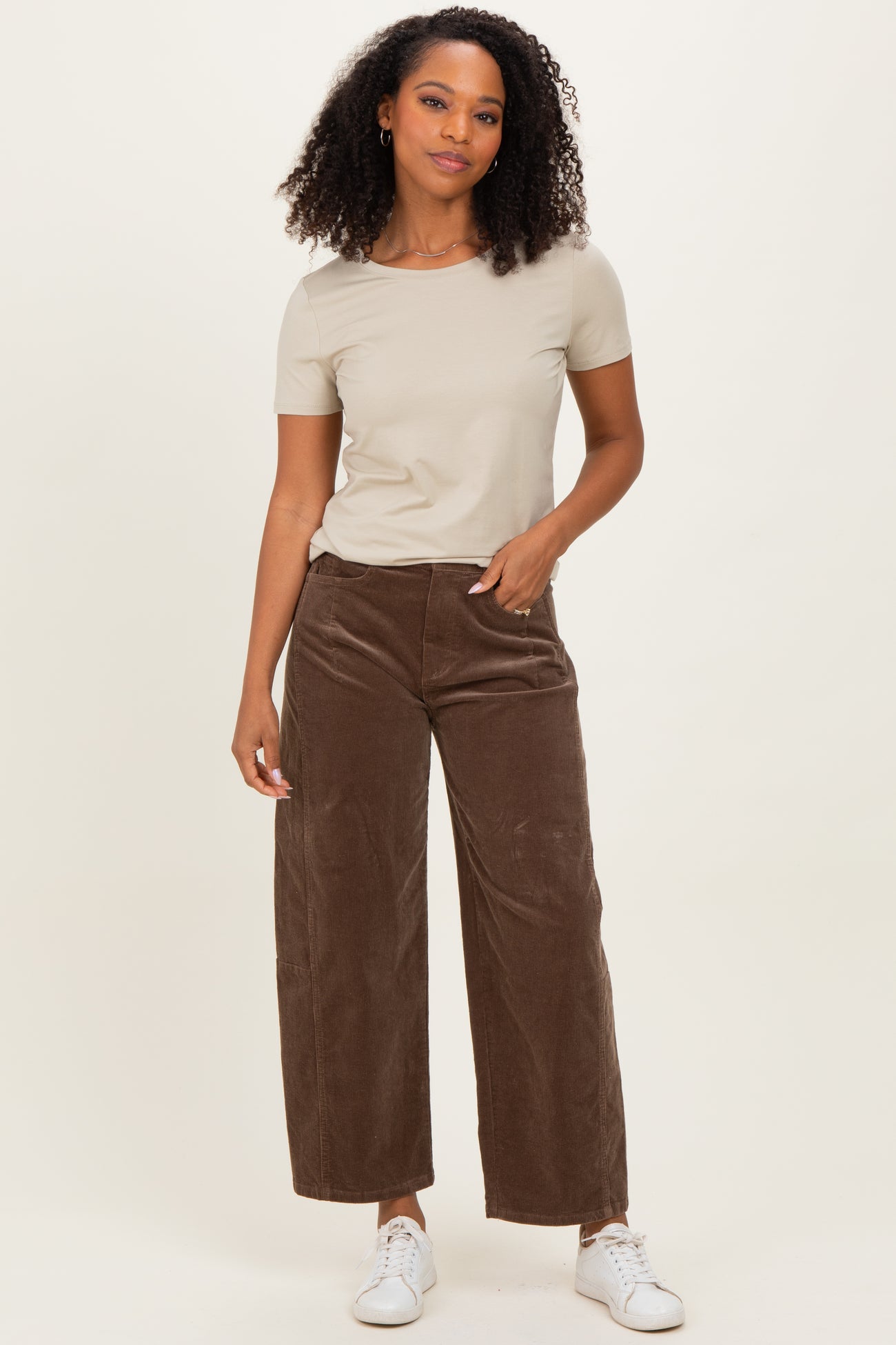 woman wearing a beige short sleeve top and brown wide-leg corduroy pants, standing with one hand on her hip