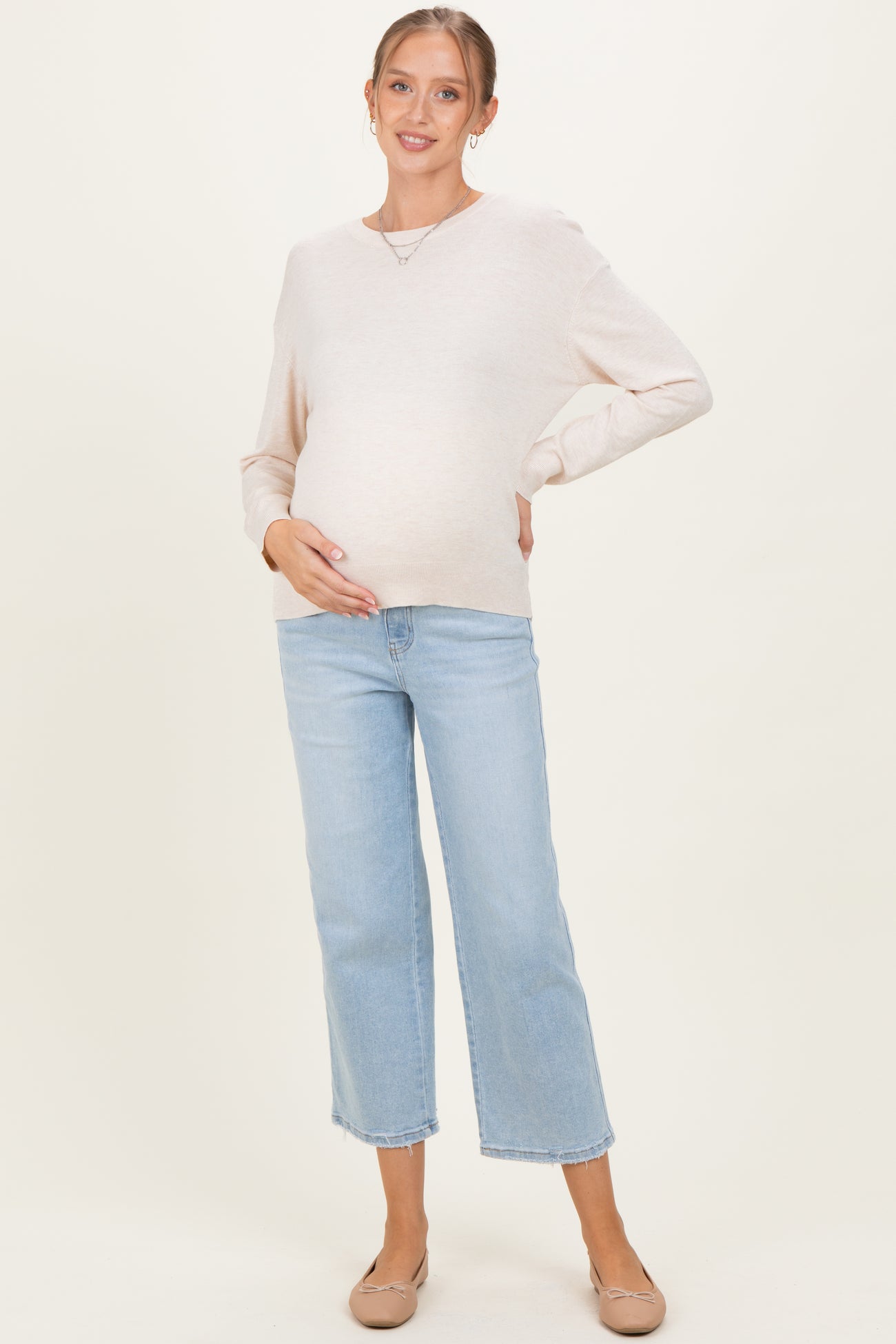woman wearing a light beige long sleeve maternity sweater and blue cropped jeans, showing her baby bump