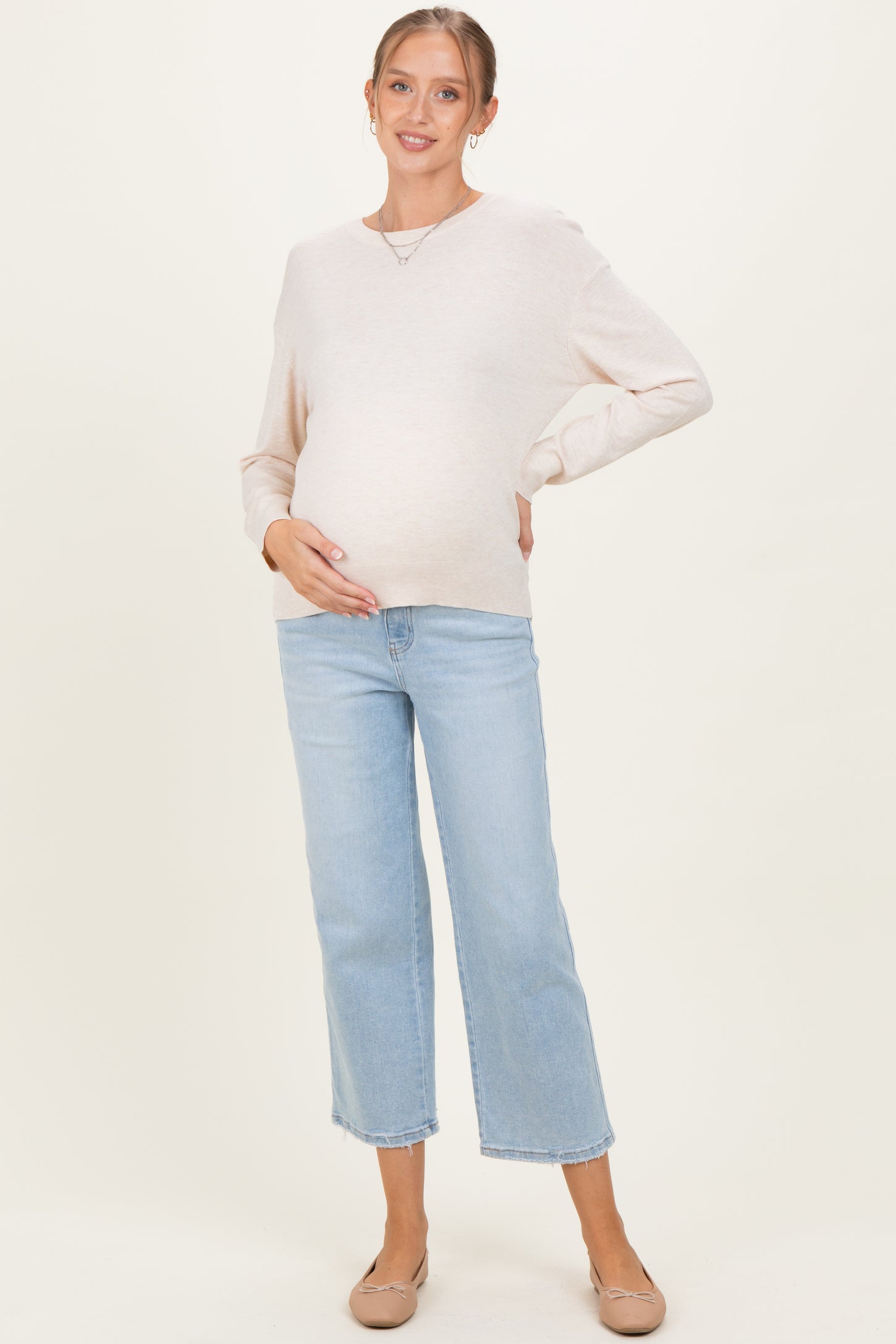 woman wearing a light beige long sleeve maternity sweater and blue cropped jeans, showing her baby bump