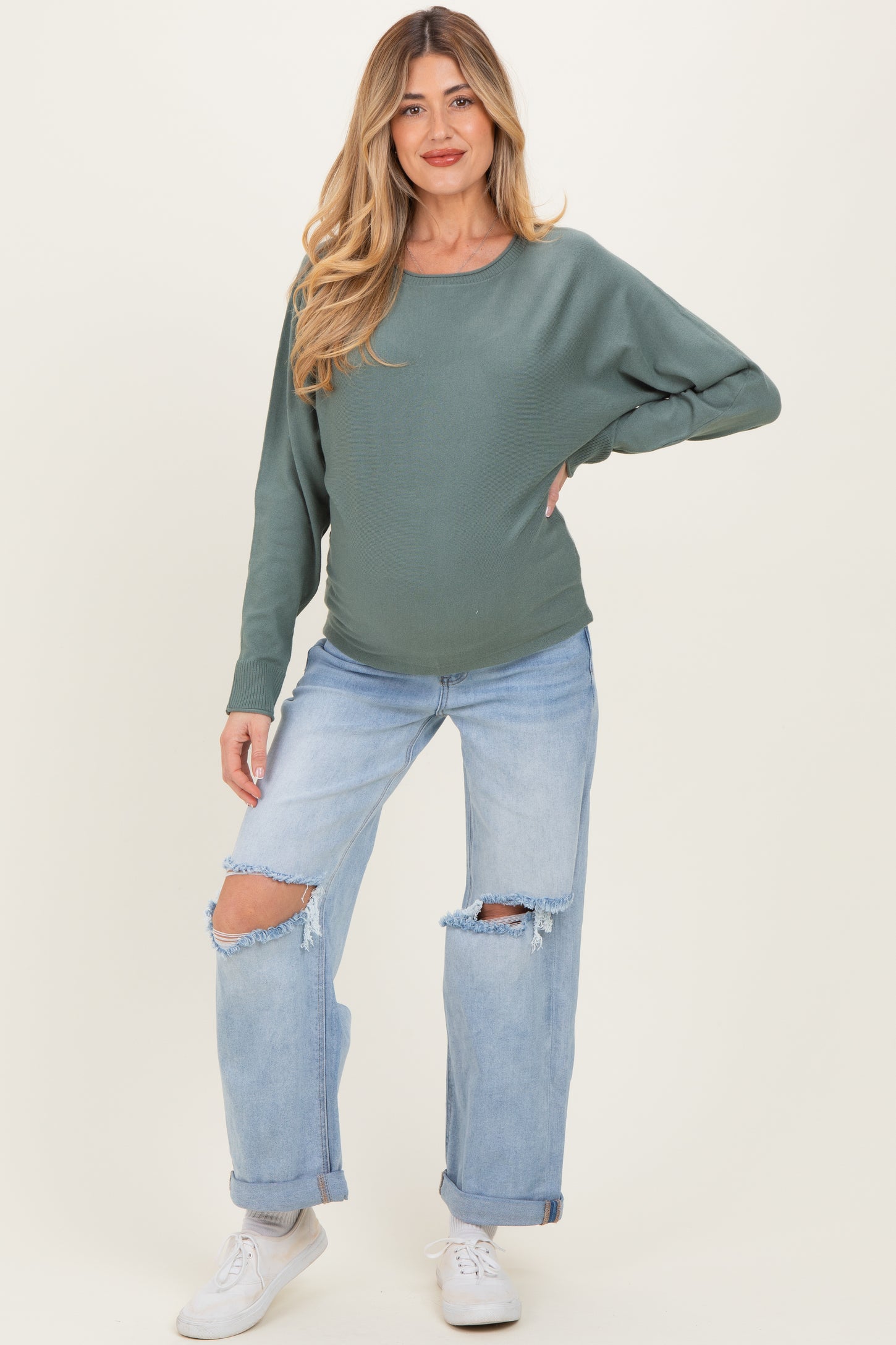 woman wearing a green long sleeve maternity top and distressed light blue jeans, showing her baby bump