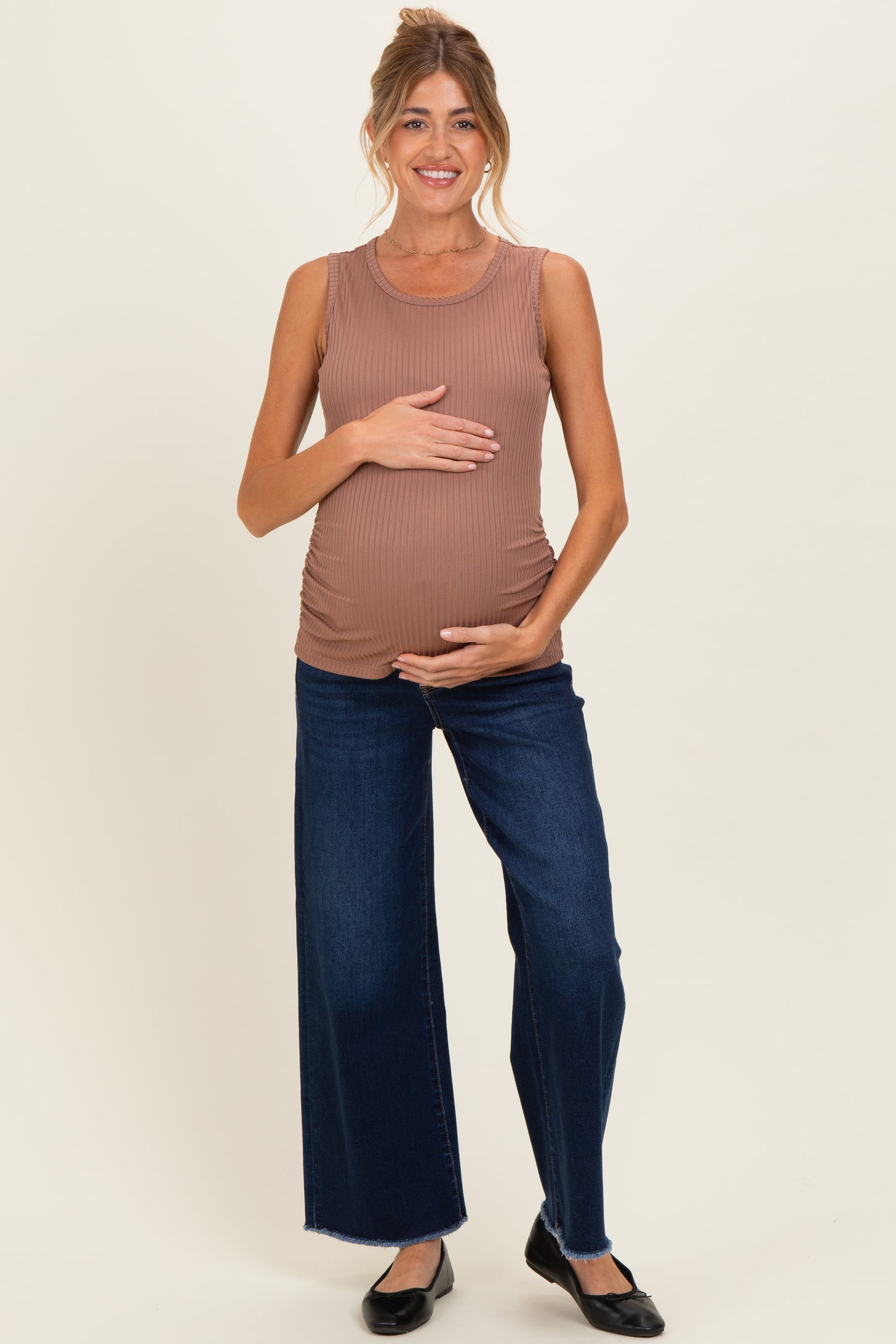 woman wearing a ribbed brown maternity tank top and dark denim wide-leg maternity jeans, holding her baby bump