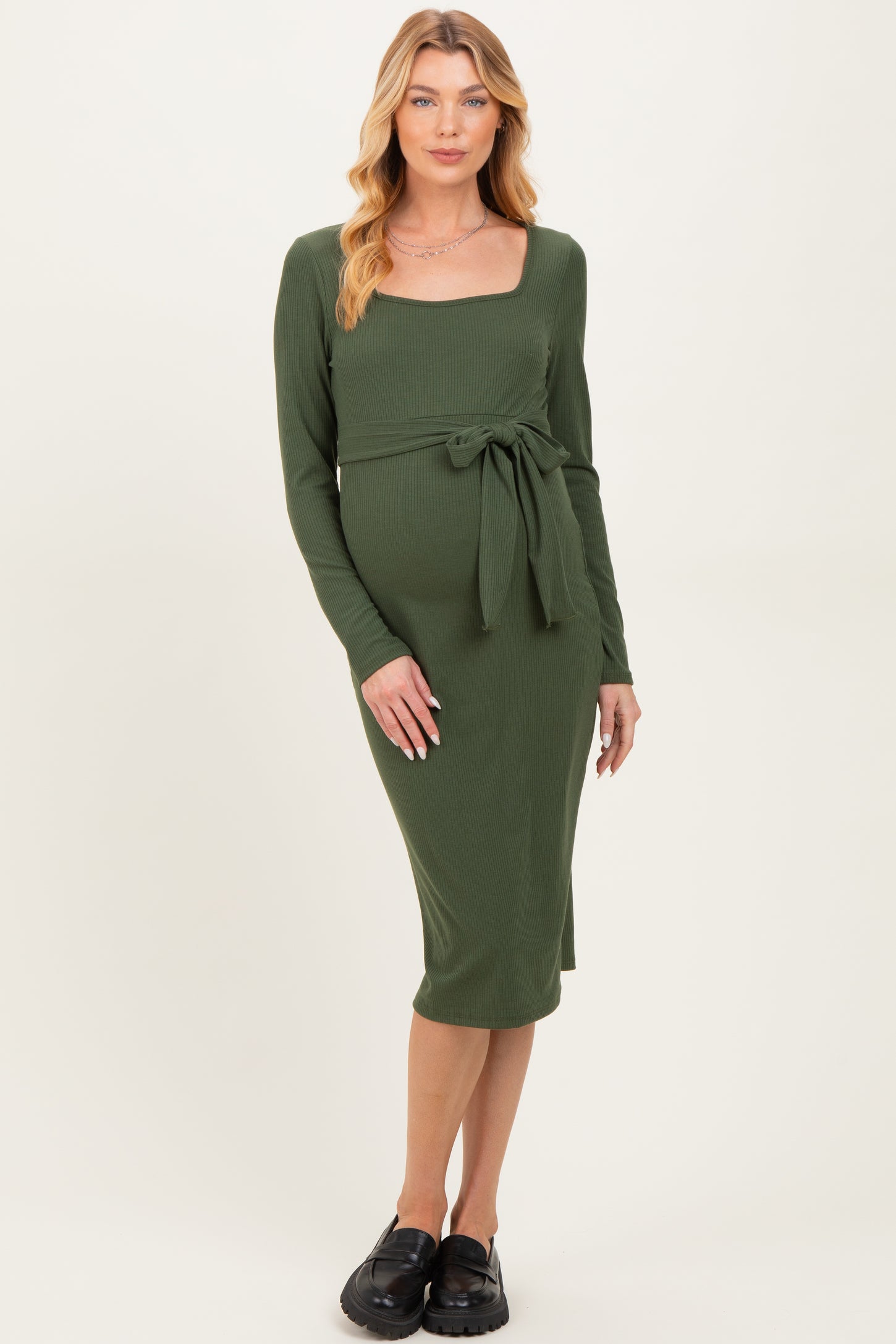 woman wearing a dark green long sleeve maternity dress with a square neckline and tie detail at the waist, showing her baby bump