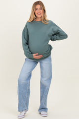 woman wearing a green oversized maternity sweatshirt and light blue wide-leg jeans, holding her baby bump