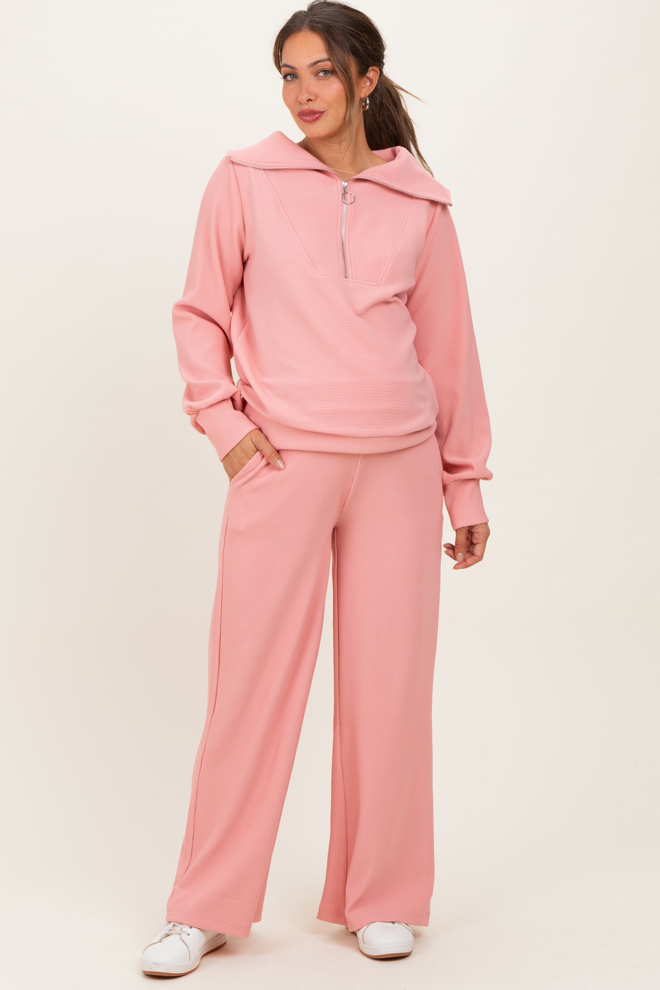 woman wearing a light pink maternity tracksuit with a zip-up hoodie and wide-leg pants, holding a baby bump