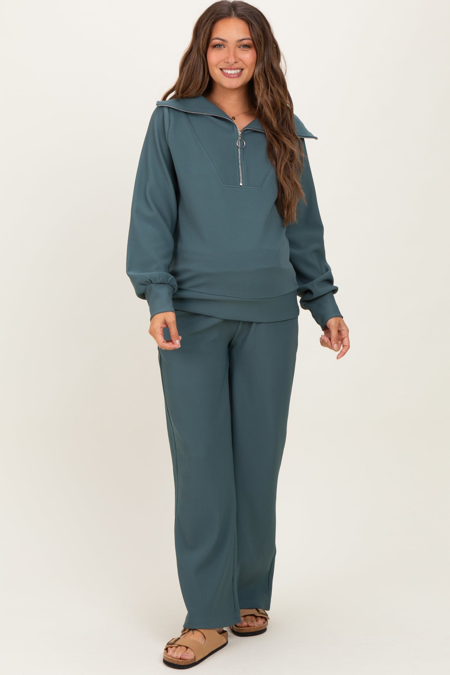 woman wearing a teal maternity tracksuit with a quarter zip pullover and wide leg pants, smiling and standing softly